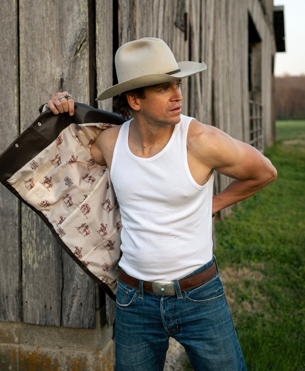 Man in a tank top holding open the H Bar C Cody jacket to show its printed lining, outside a barn.