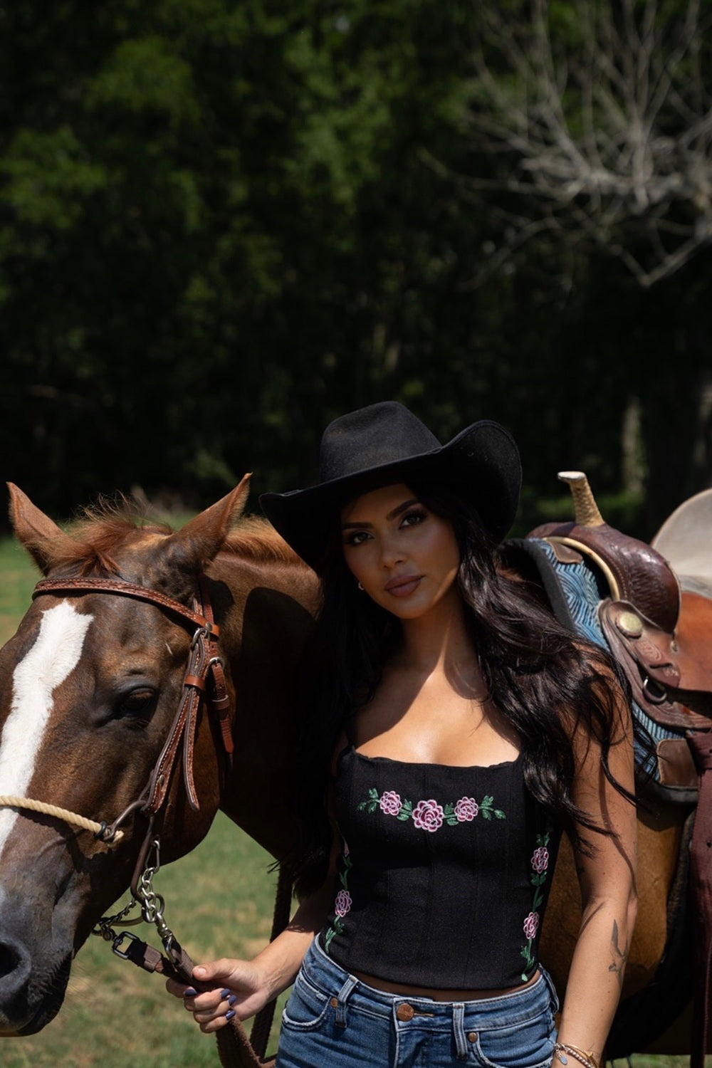 Woman in a black cowboy hat wearing the H Bar C Canterbury black corset with jeans, standing beside a horse.