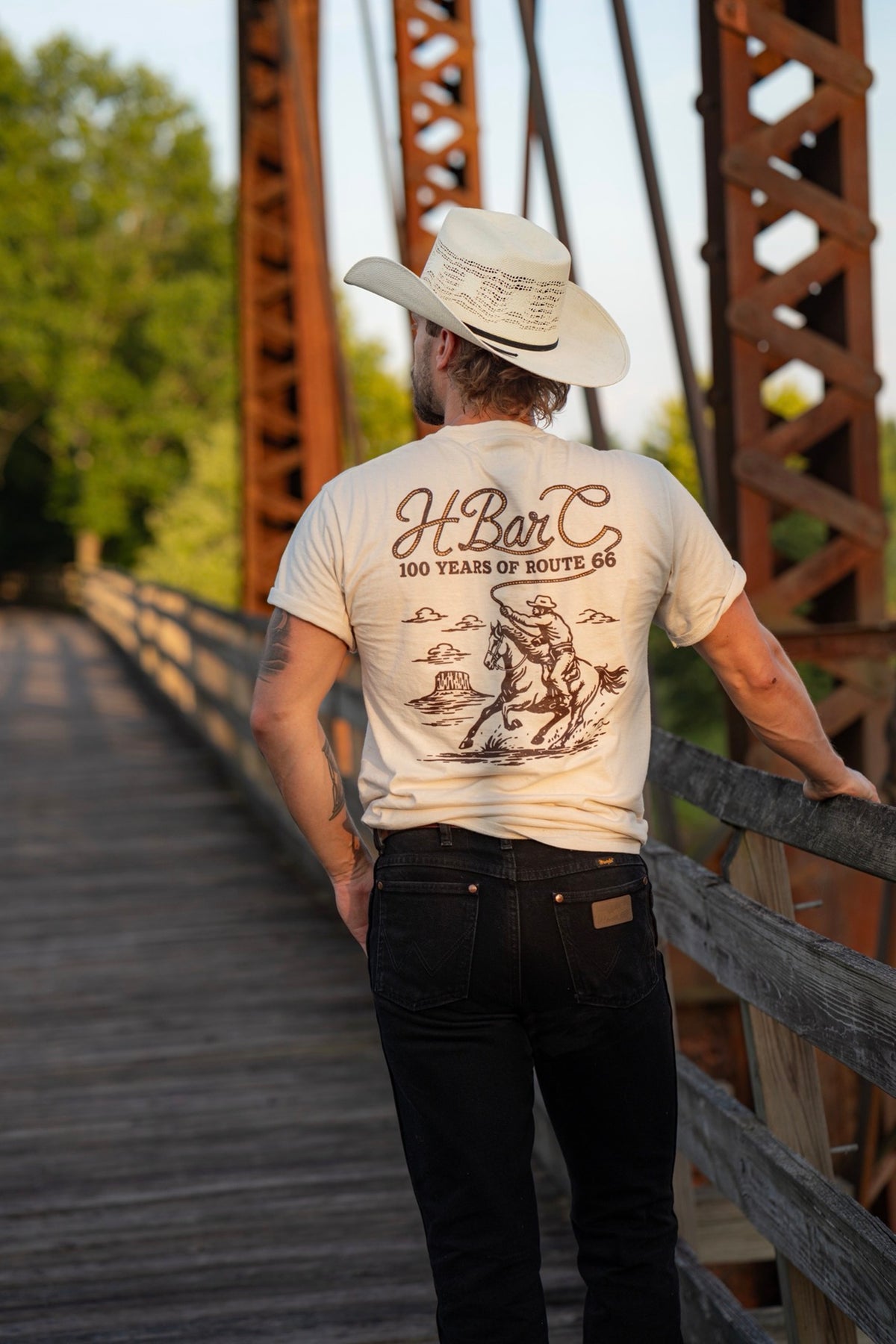 Man in a cowboy hat showing the back graphic of the H Bar C Route 66 Centennial Tulsa Rope tee on an old truss bridge.