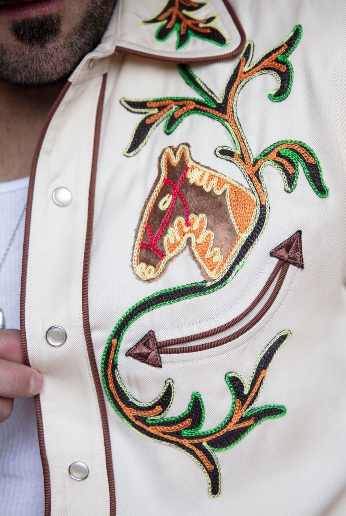 Chest detail of the H Bar C Calabasas off-white shirt with a chain stitch horse head, arrow, and green vine embroidery.