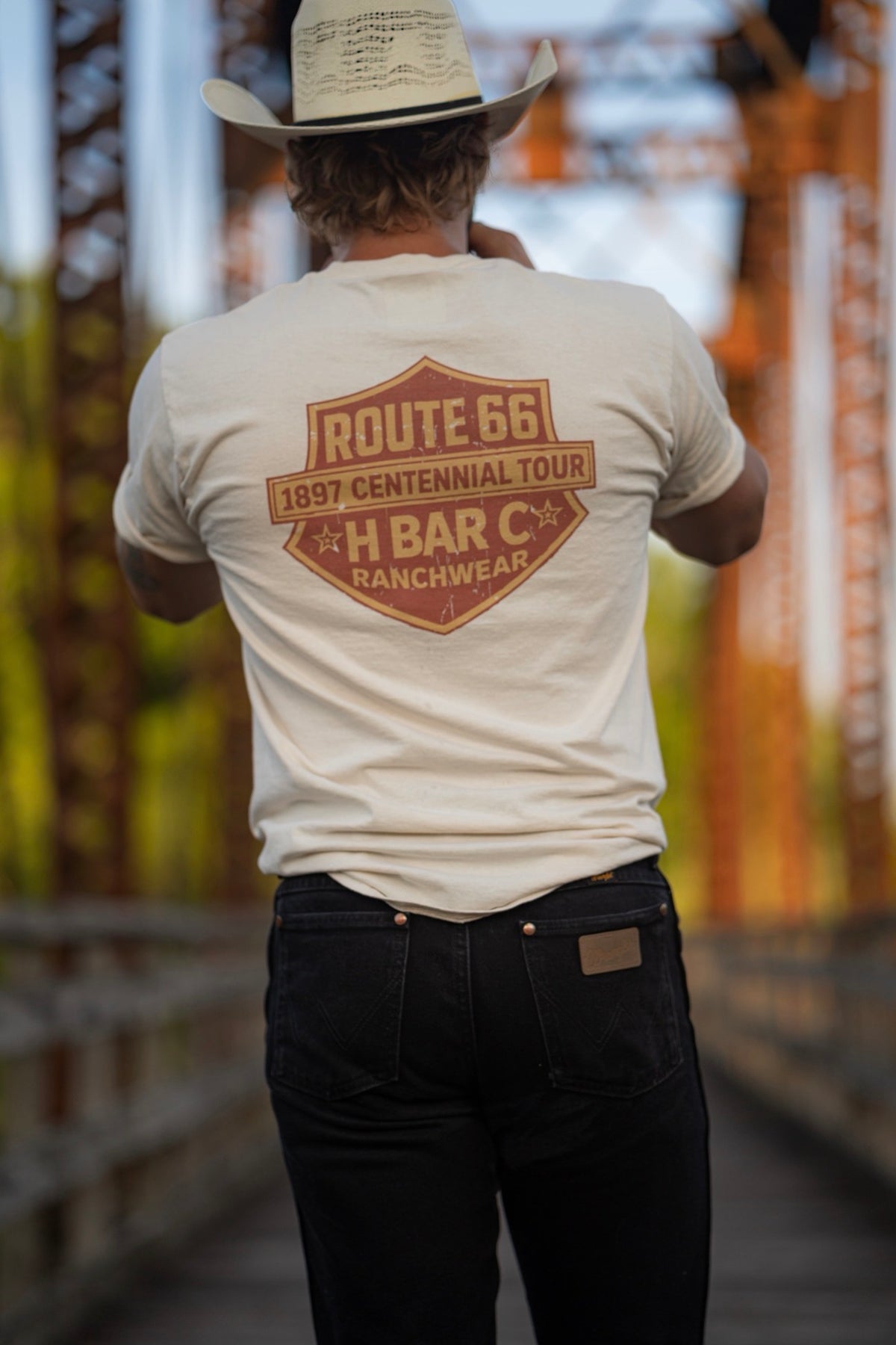 Back of the H Bar C Tulsa Shield T-shirt with an orange Route 66 shield graphic, worn on a rusted iron bridge.