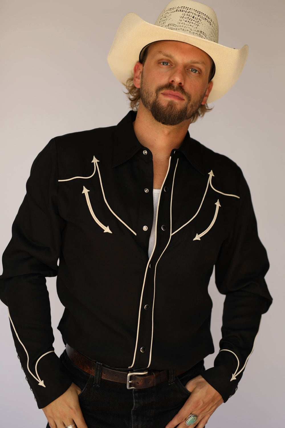 Man in a cream cowboy hat wearing the H Bar C Reno black shirt with off-white arrow piping, a Man in Black tribute, with dark jeans.