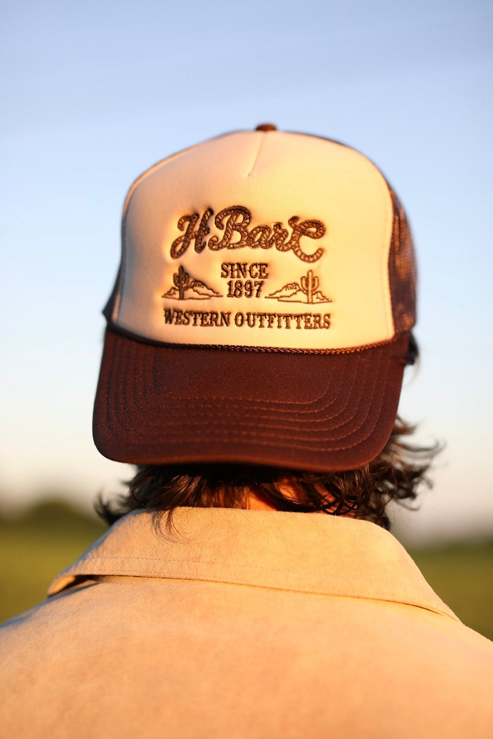 H Bar C Western Outfitters trucker hat in tan with brown rope script logo and brown mesh, worn outdoors at sunset.