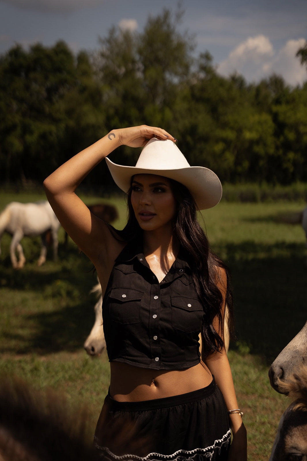 Woman in a white cowboy hat wearing the H Bar C Hampton halter top in black in a pasture with horses.