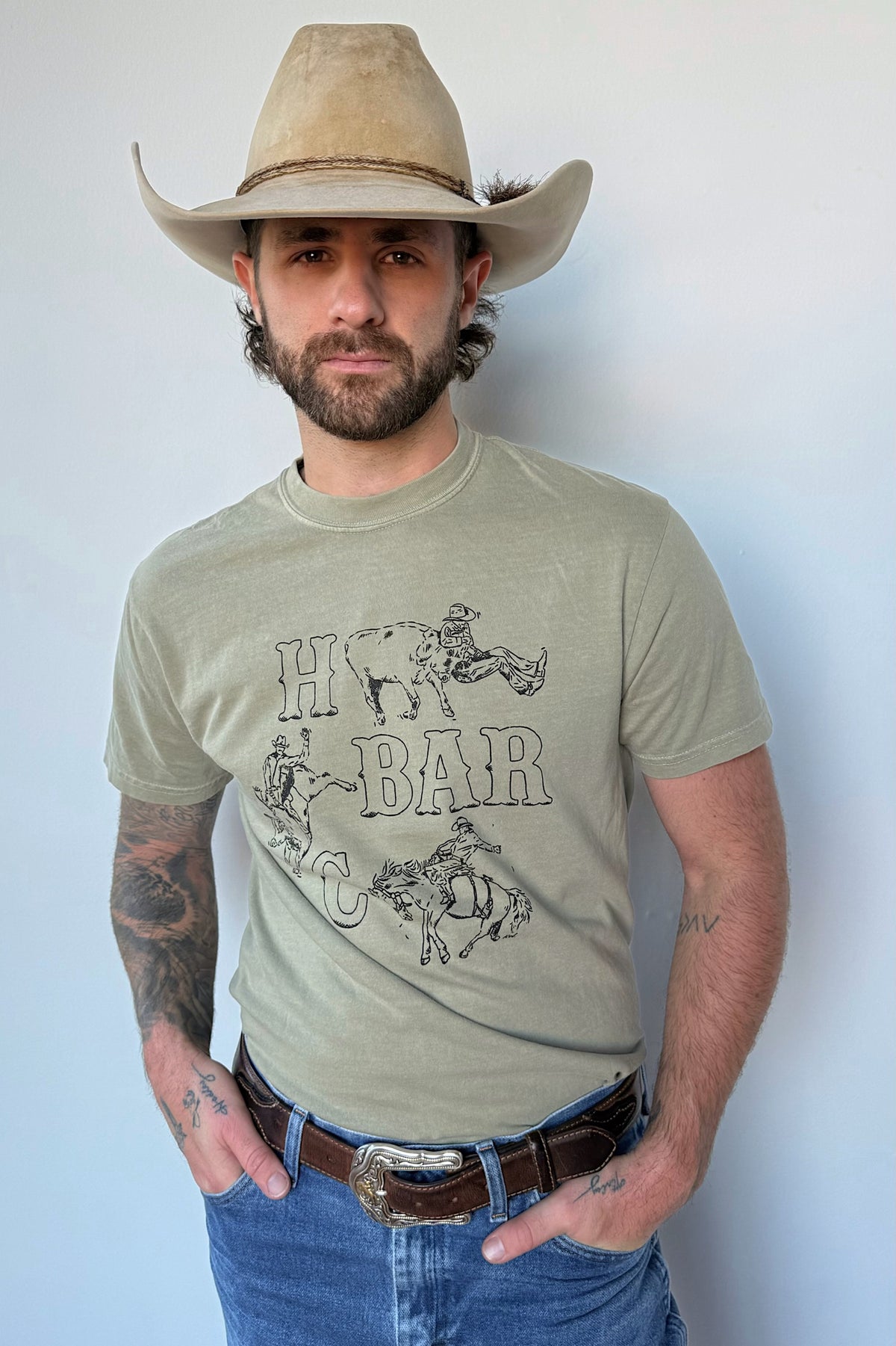 Man in a straw cowboy hat wearing the H Bar C Busted Spur tee in green, a Kate Snake Art collaboration with a hand-drawn bronc rider.