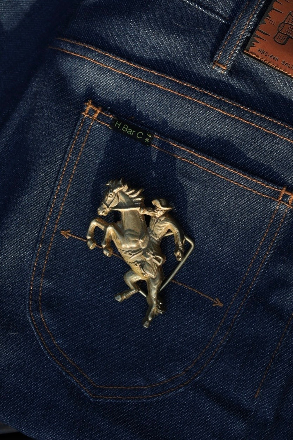 H Bar C gold bucking horse and rider belt buckle on a dark denim back pocket.
