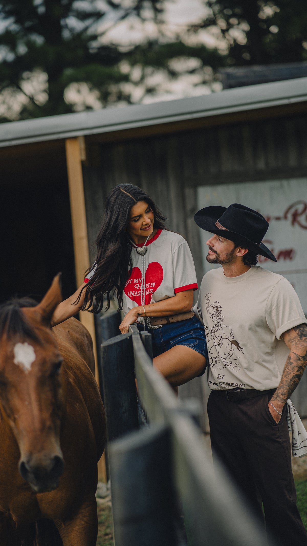 Couple by a horse at a ranch fence, she wears the H Bar C x Rustler Left My Heart in Nashville ringer tee.