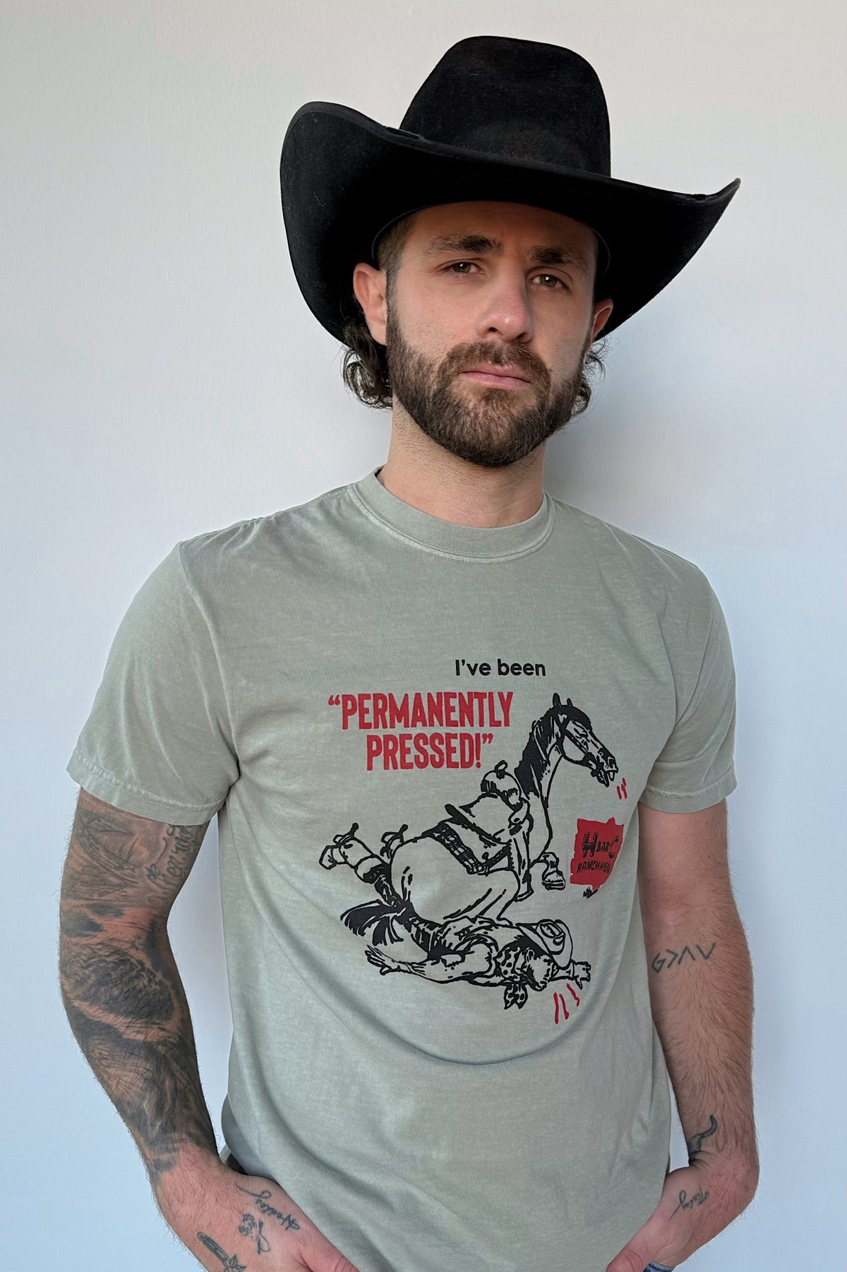 Man in a black cowboy hat wearing the H Bar C Dry Gulch tee in olive with a vintage Permanently Pressed bucking horse graphic.
