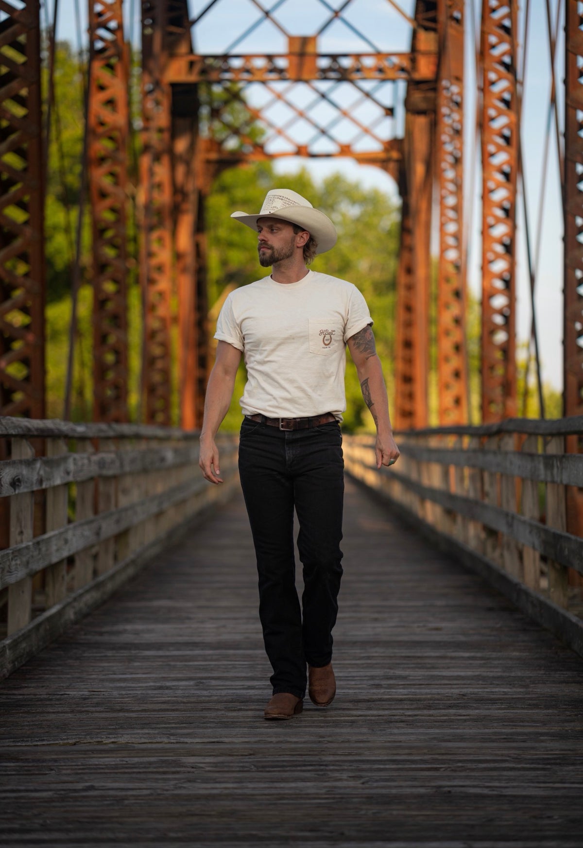 Man walking across a vintage iron bridge wearing the H Bar C Tulsa Rope Route 66 tee with black jeans and cowboy hat.