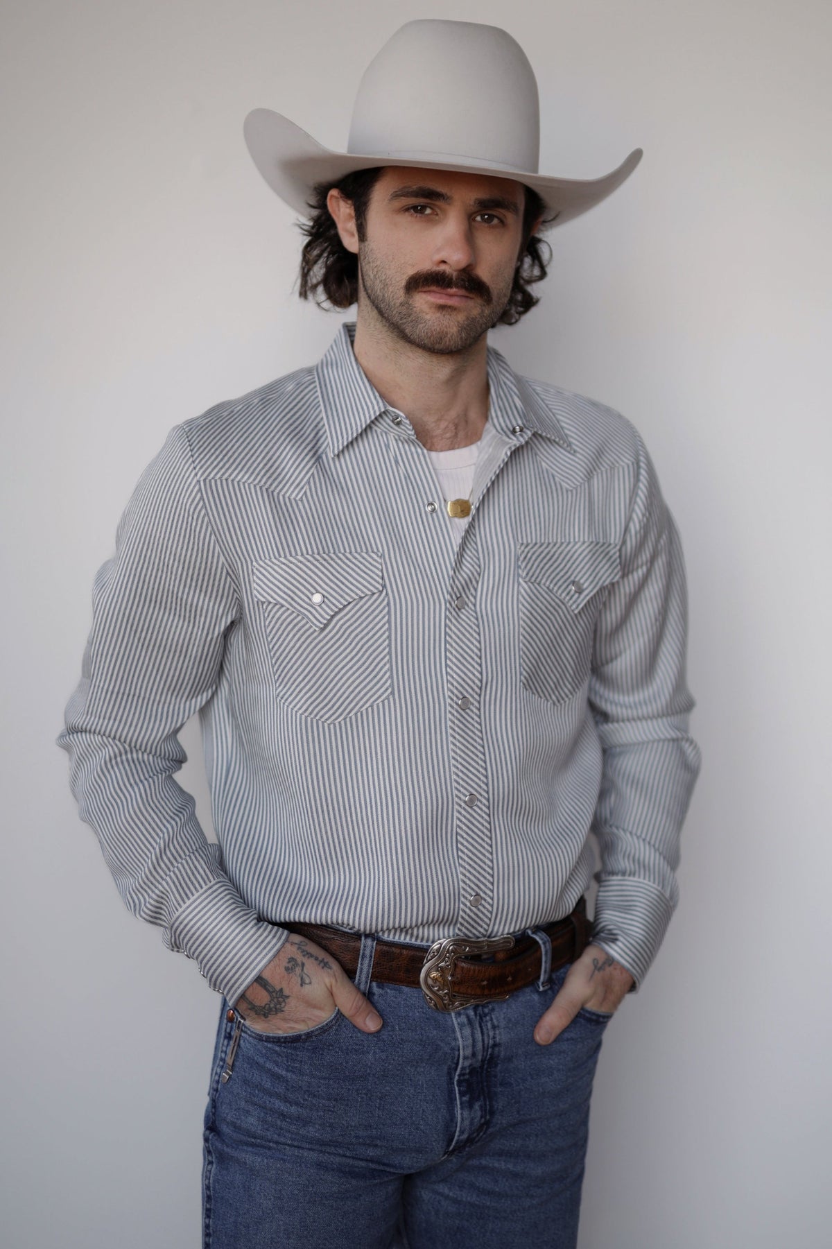 Man in a cream cowboy hat wearing the H Bar C Folsom shirt, a blue and white pinstripe pearl snap western shirt, with jeans.