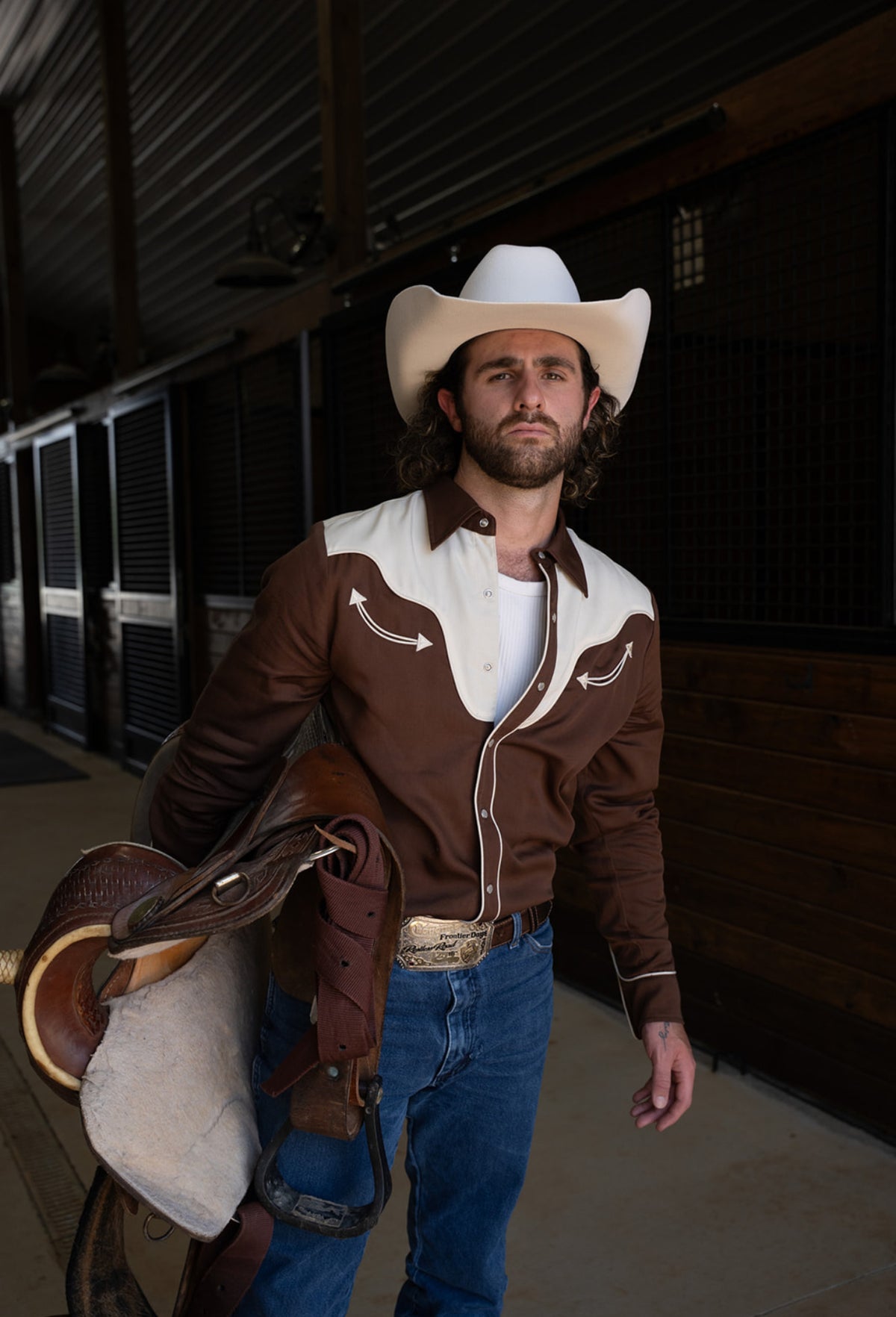 Man carrying a saddle in a barn wearing the H Bar C San Juan chocolate two-tone western shirt and jeans.
