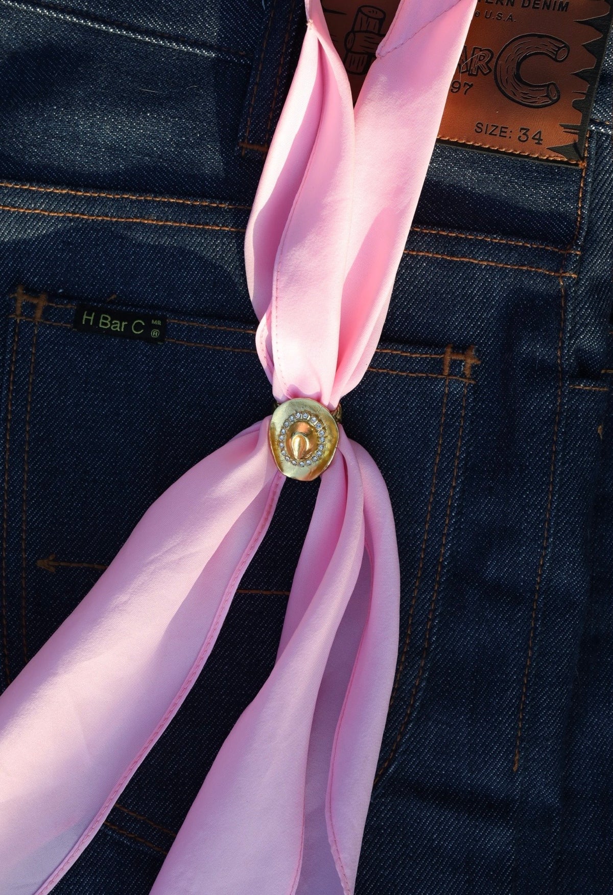 Pink wild rag threaded through the H Bar C gold cowboy hat scarf slide on denim.