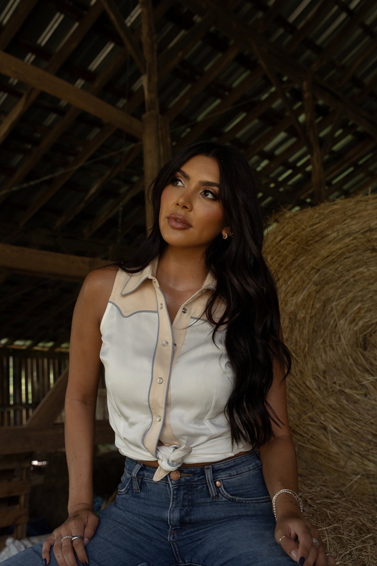Woman in a barn wearing the H Bar C Atlanta women's sleeveless shirt in white and cream with jeans.