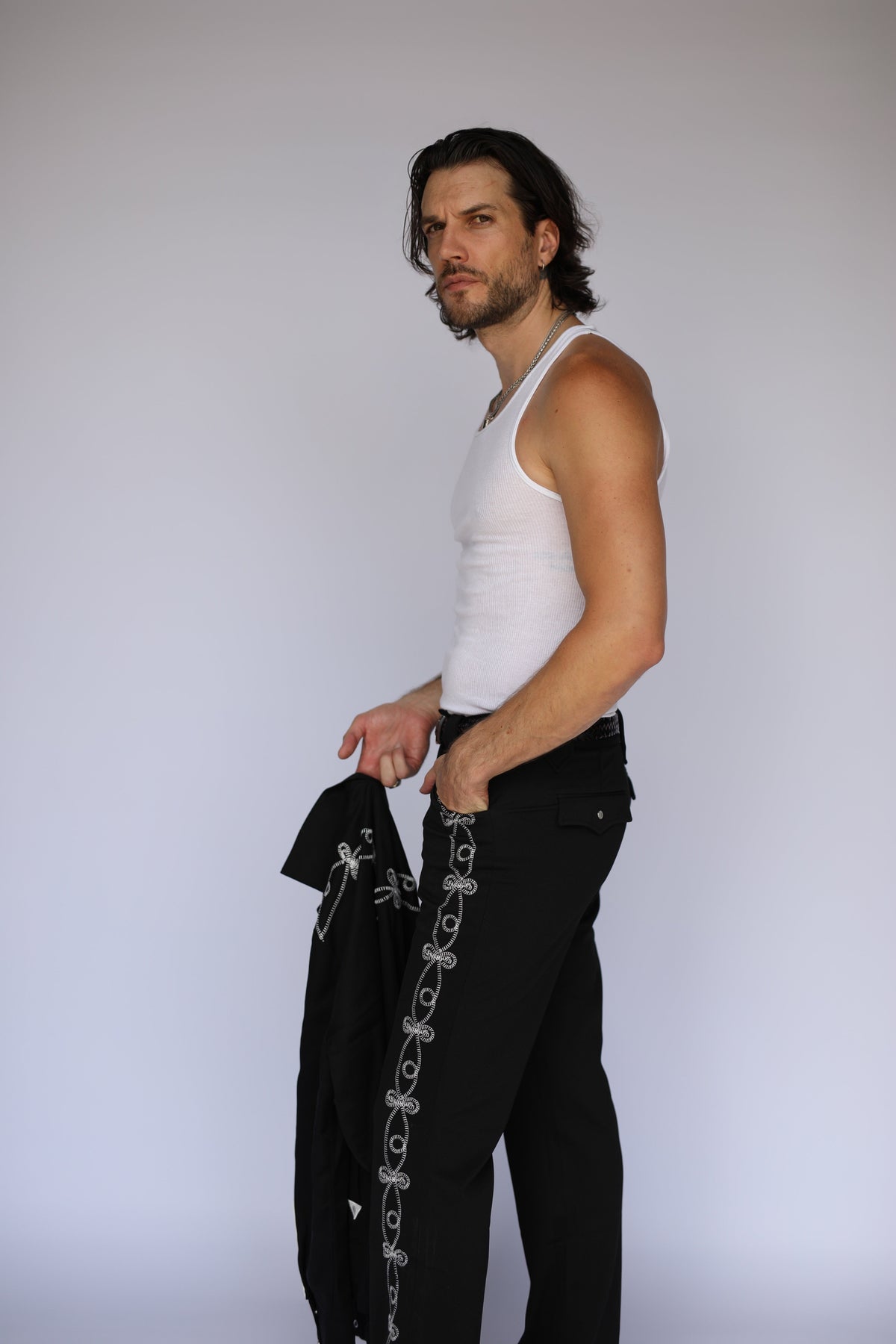 Side view of the H Bar C Bolero Pants El Patrón in black with soutache braid down the leg, held jacket in hand.