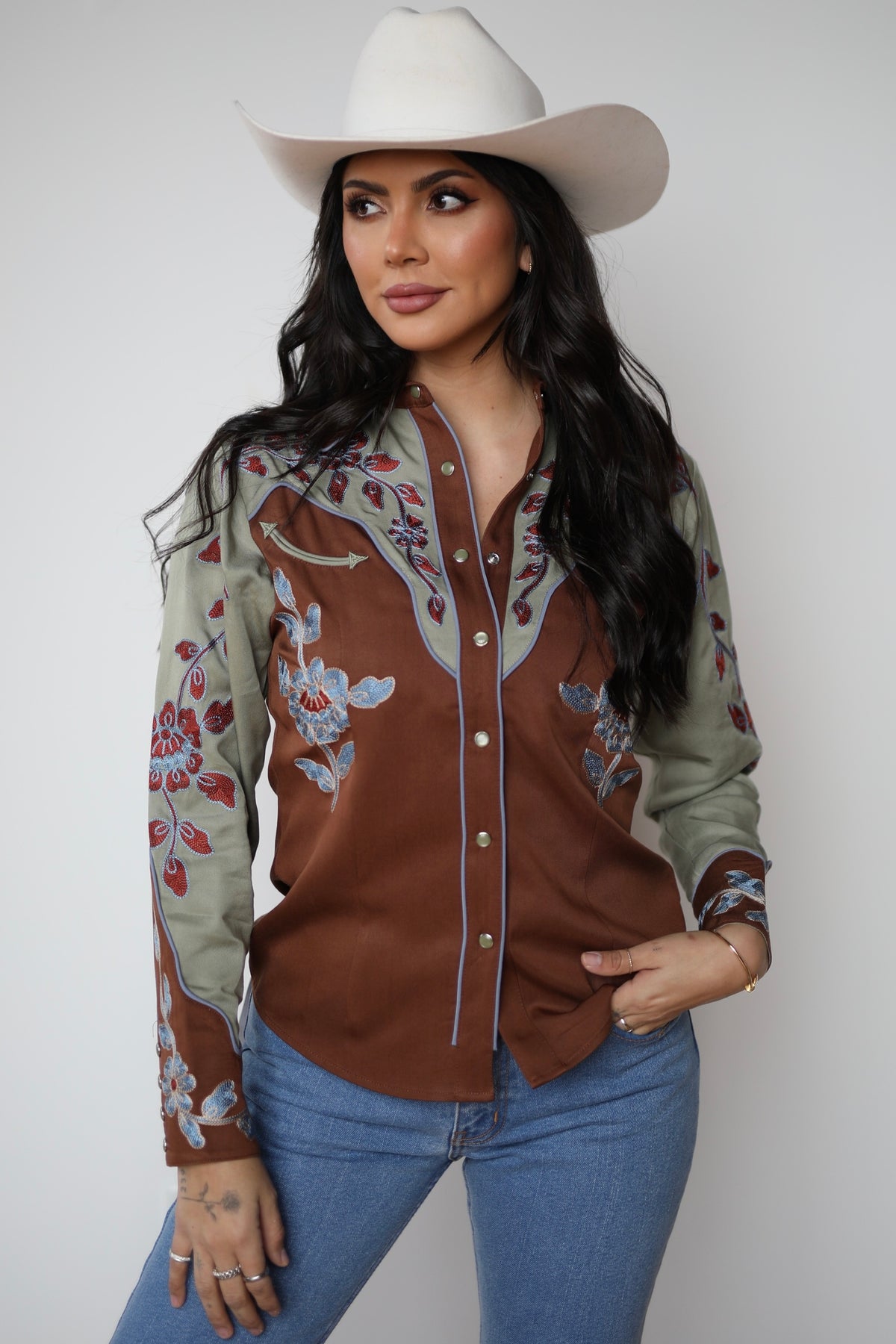 Woman in a white cowboy hat wearing the H Bar C San Antonio women's shirt in brown with sage yokes and sleeves and red floral embroidery.