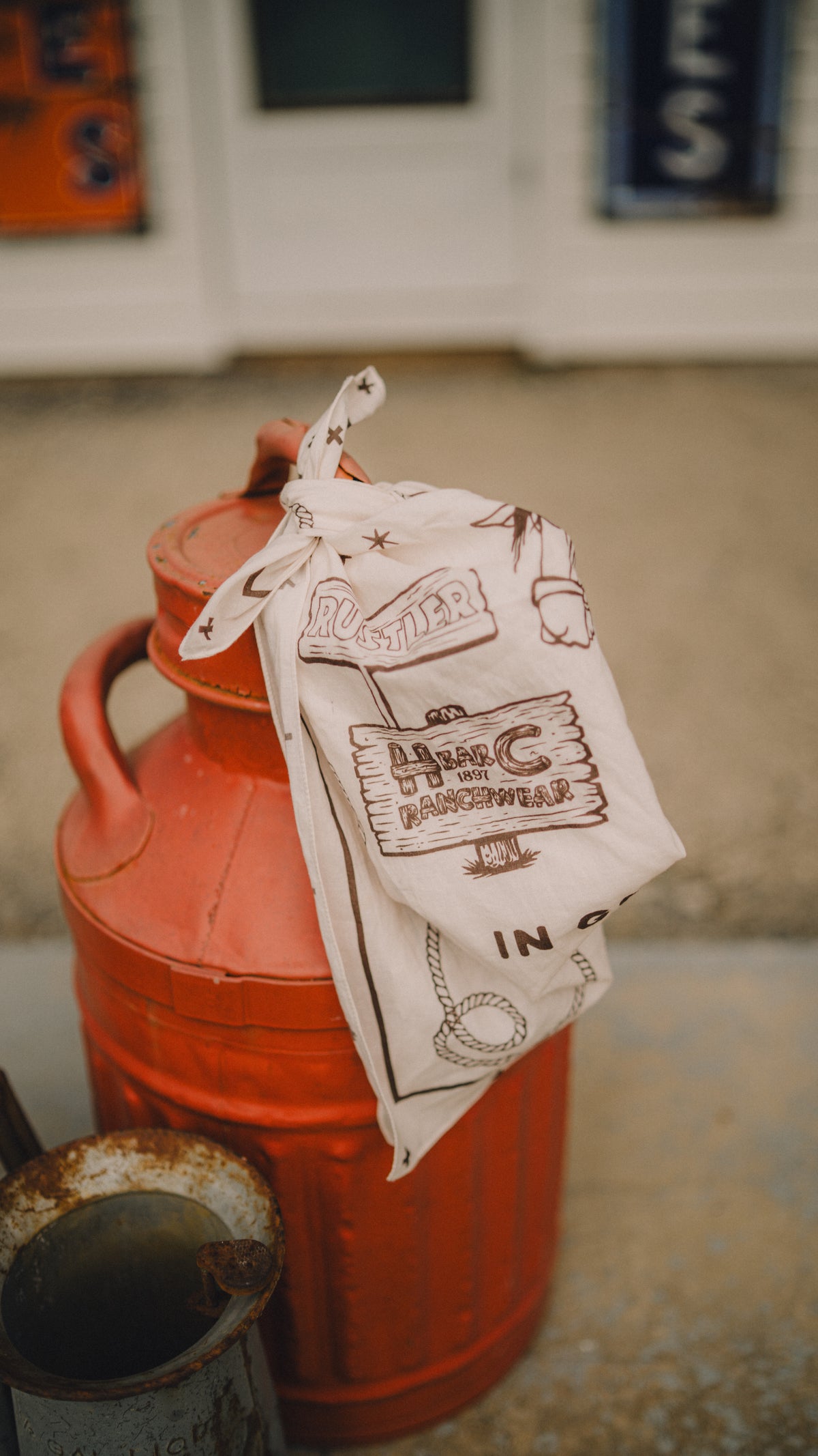 H Bar C x Rustler western bandana draped over a vintage red oil can.