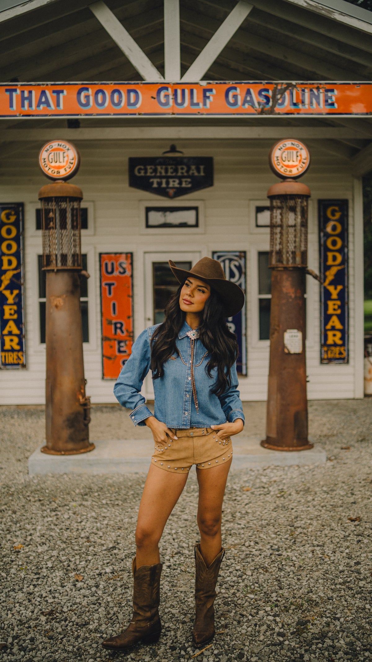 Woman wearing the Rustler x H Bar C denim cropped shirt and tan pants in front of a vintage Gulf gas station.