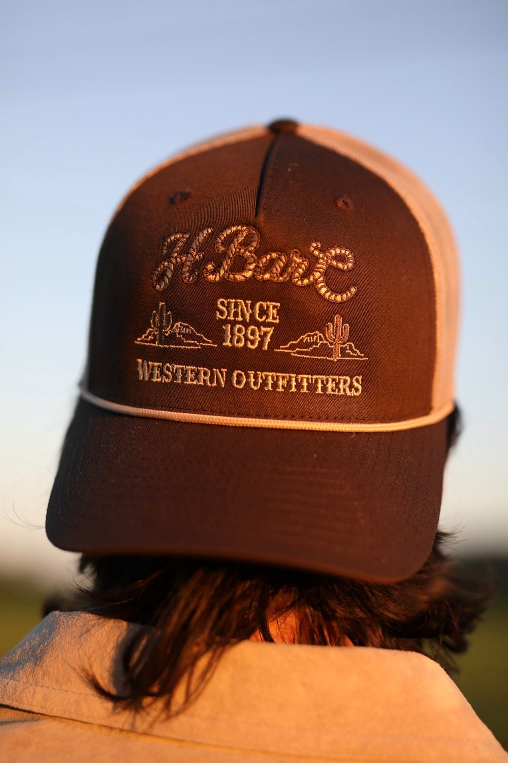 H Bar C Western Outfitters trucker hat in brown with cream rope script and Since 1897, worn outdoors.