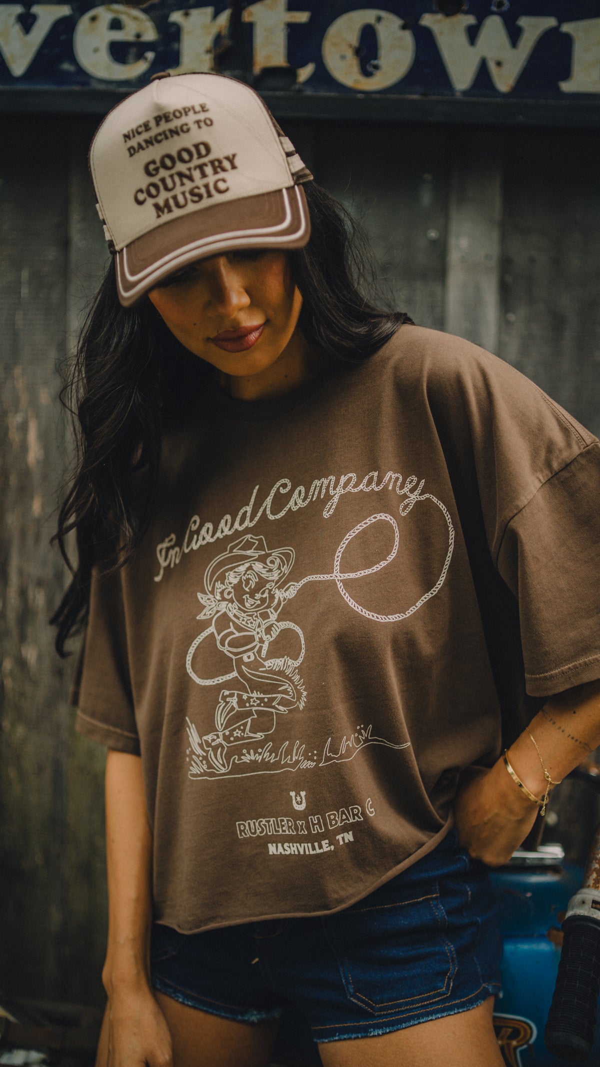 Woman in a trucker cap wearing the H Bar C x Rustler In Good Company women's crop tee.