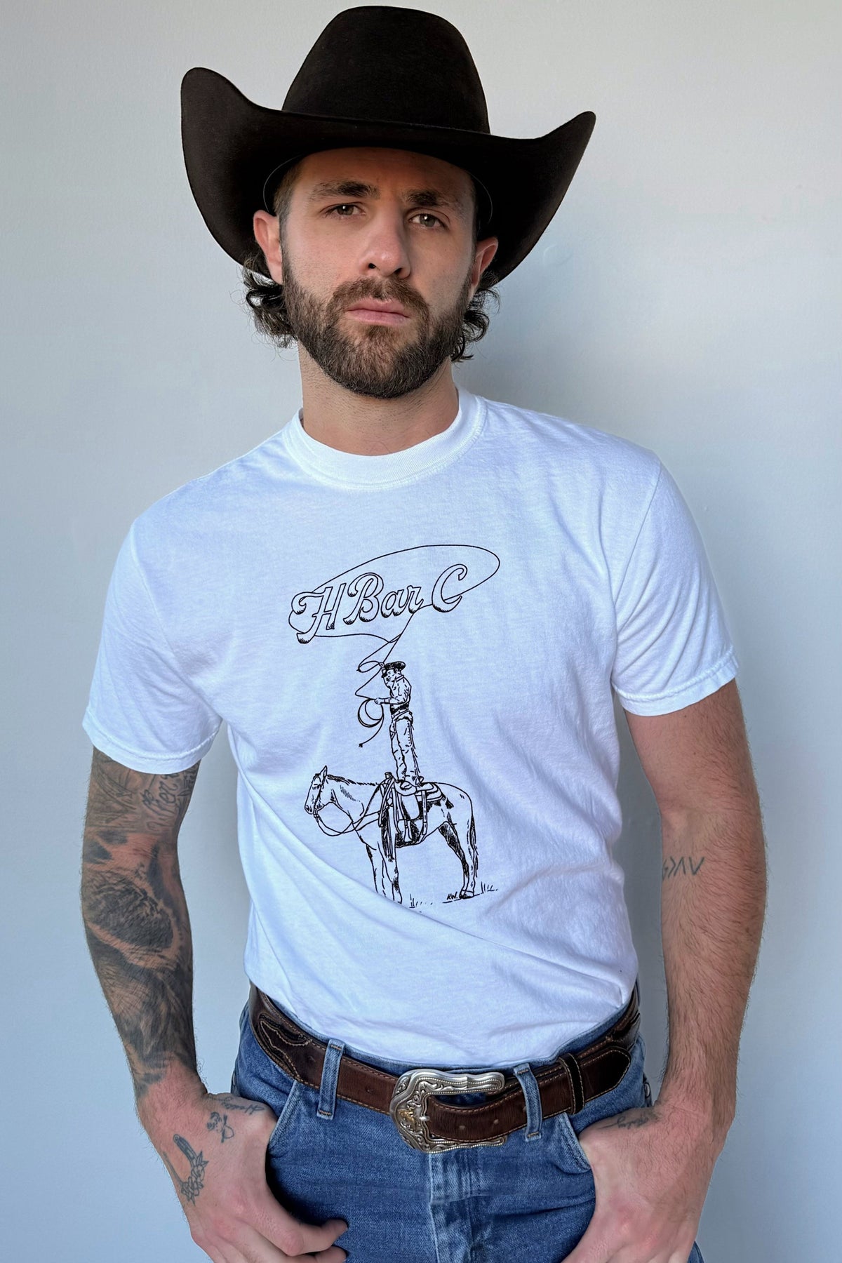 Man in a black cowboy hat wearing the H Bar C Tall Saddle white tee, a Kate Snake Art collaboration with a cowboy standing in the saddle.