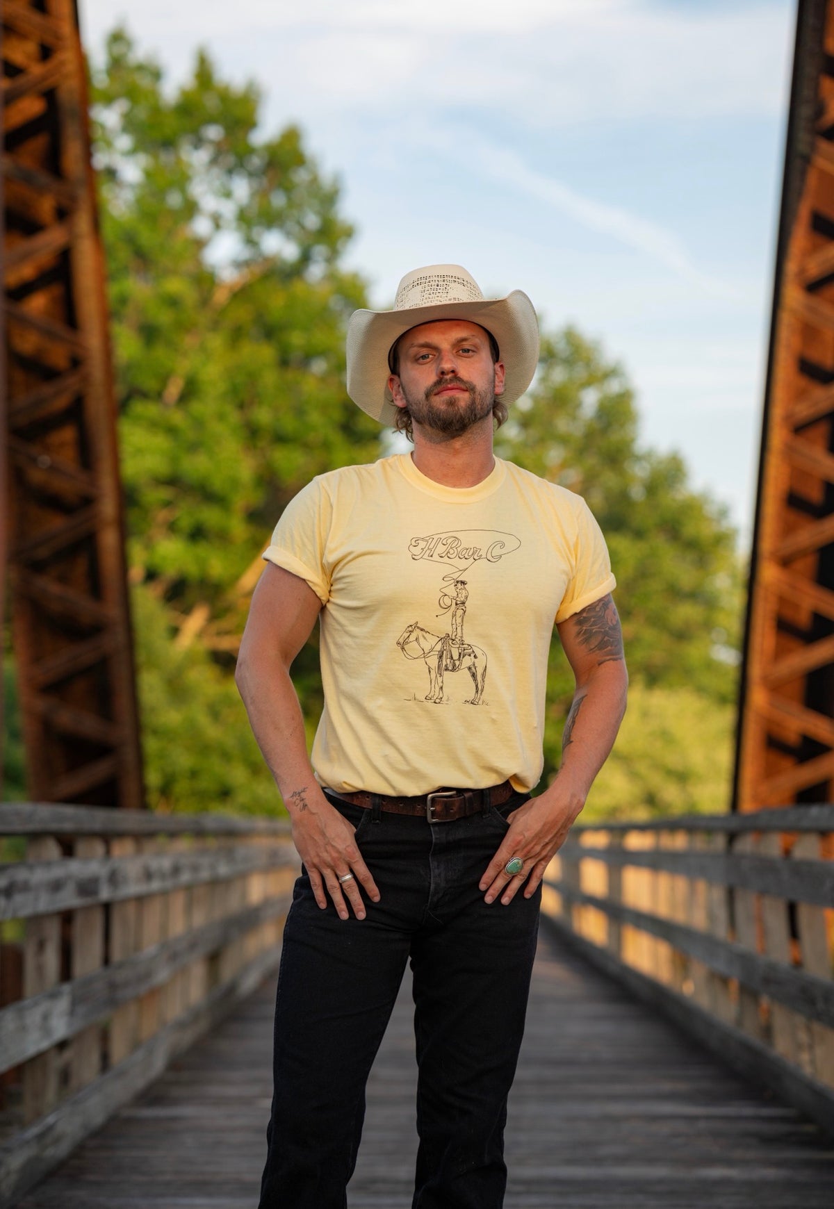 Full length view of the H Bar C Tall Saddle yellow tee on a vintage truss bridge.