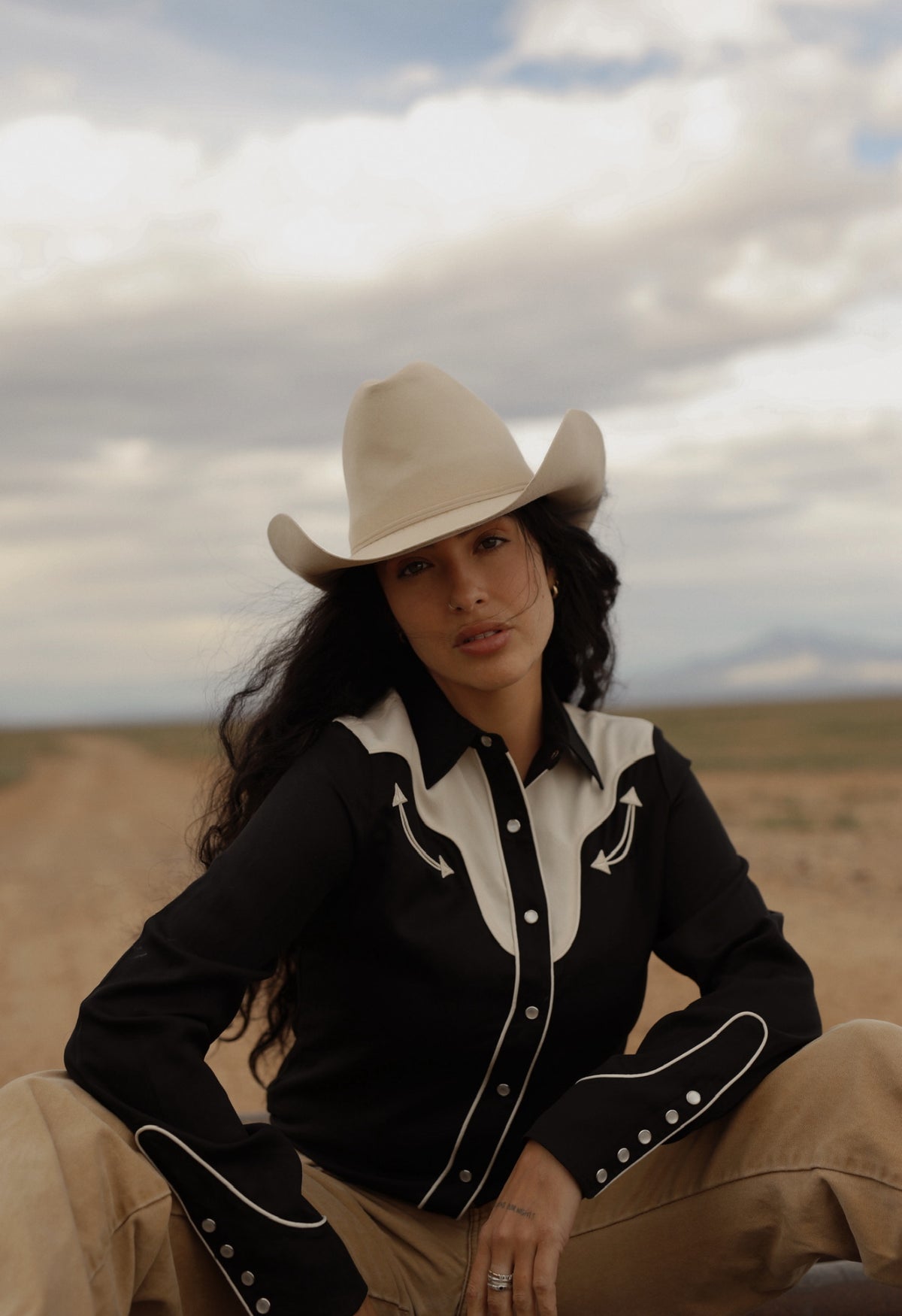 Woman in a cowboy hat seated in the desert wearing the H Bar C San Juan black and off white women's shirt.