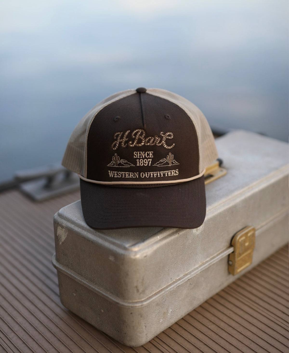 H Bar C Western Outfitters brown rope trucker hat resting on a vintage tackle box.