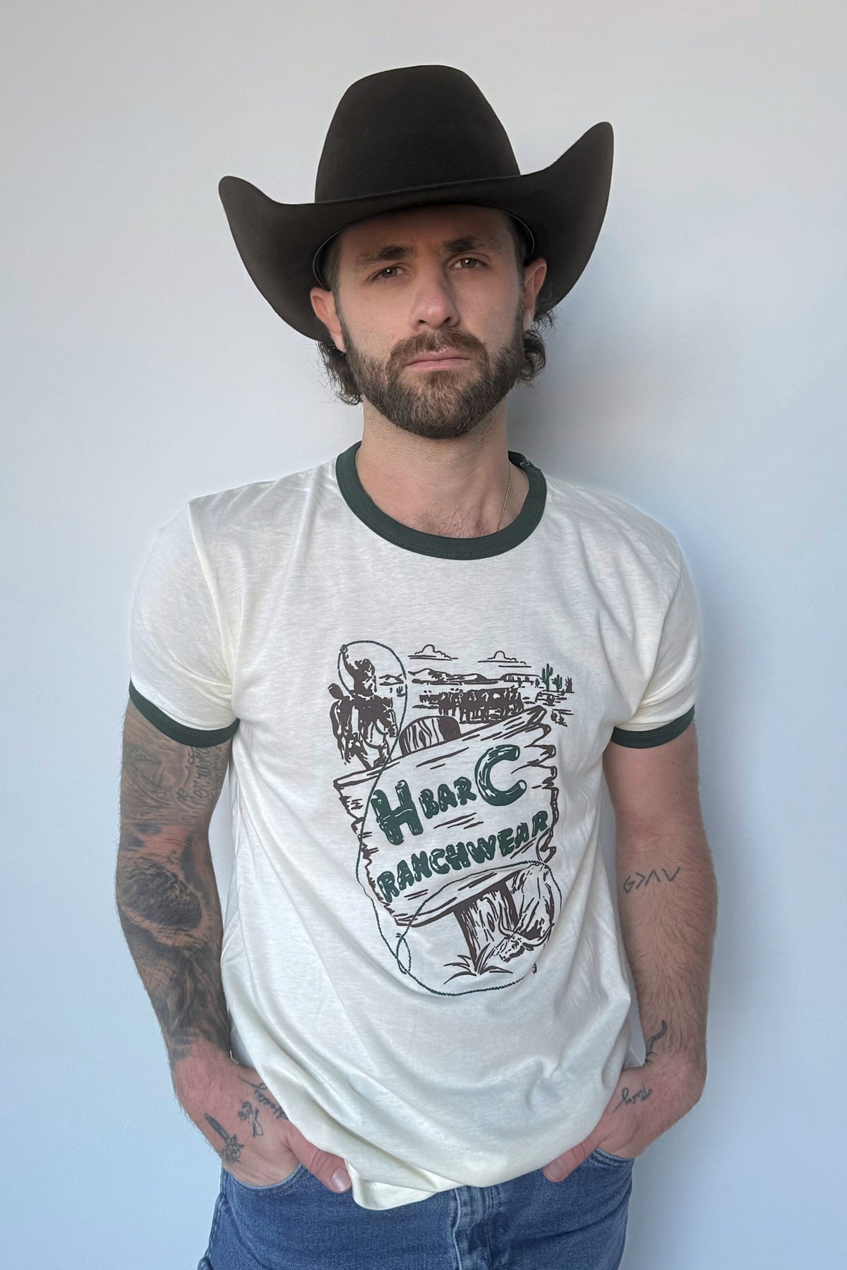 Man in a black cowboy hat wearing the H Bar C Post Canyon ringer tee in off white with green trim and vintage lasso cowboy graphic.