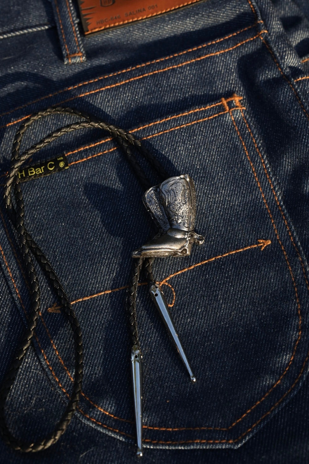 H Bar C silver cowboy boot bolo tie laid on an indigo jean back pocket.
