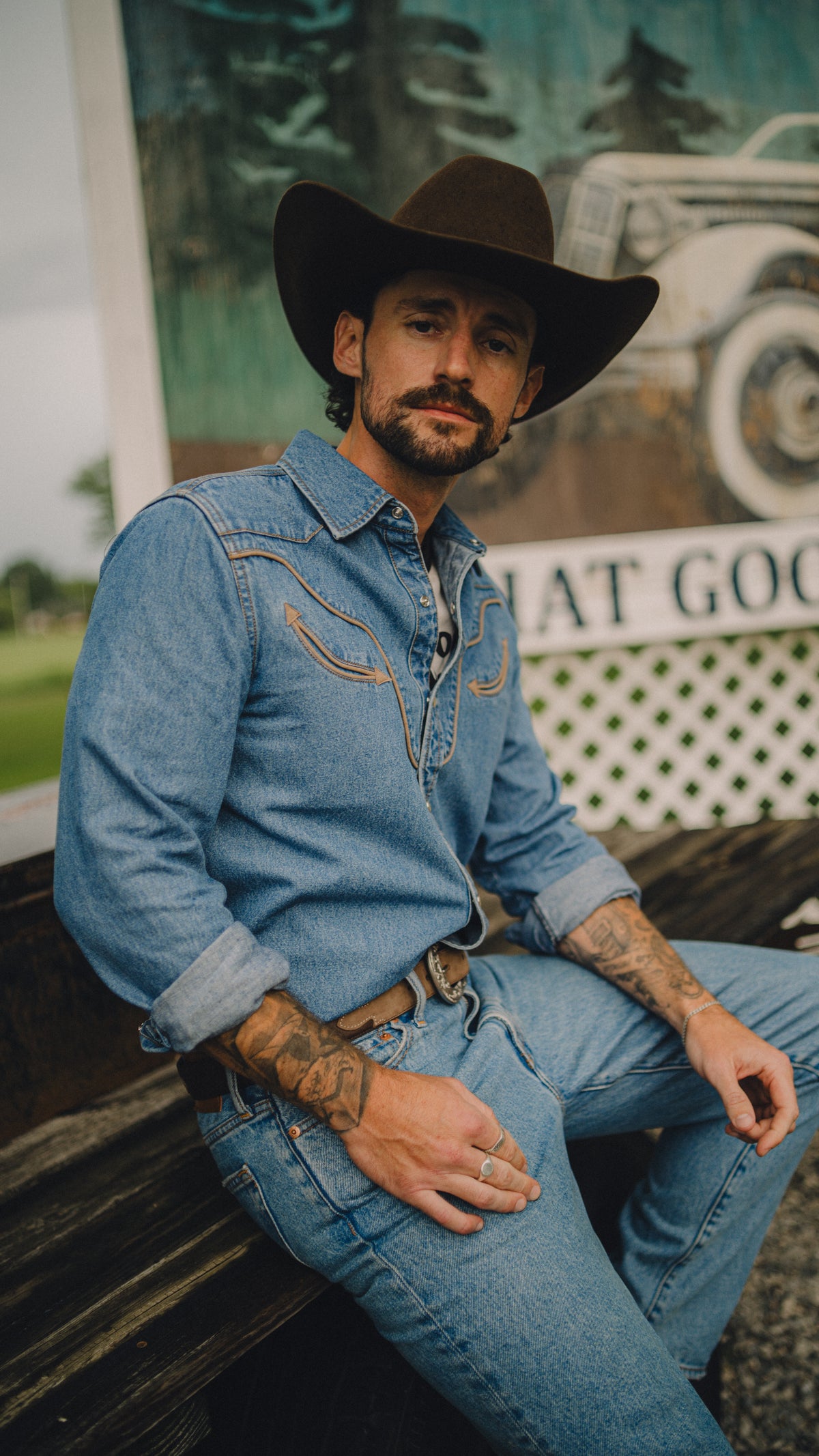 Man in a black cowboy hat sitting on a vintage car wearing the Rustler x H Bar C men's denim western shirt and jeans.