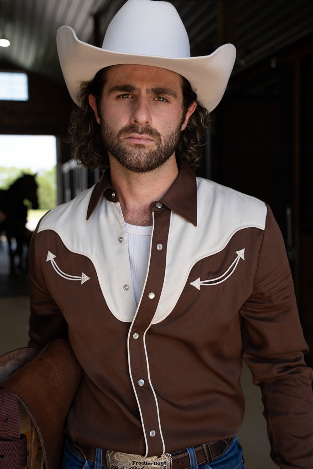 Man in a cream cowboy hat in a horse barn wearing the H Bar C San Juan men's shirt in chocolate with an off-white yoke and arrows.