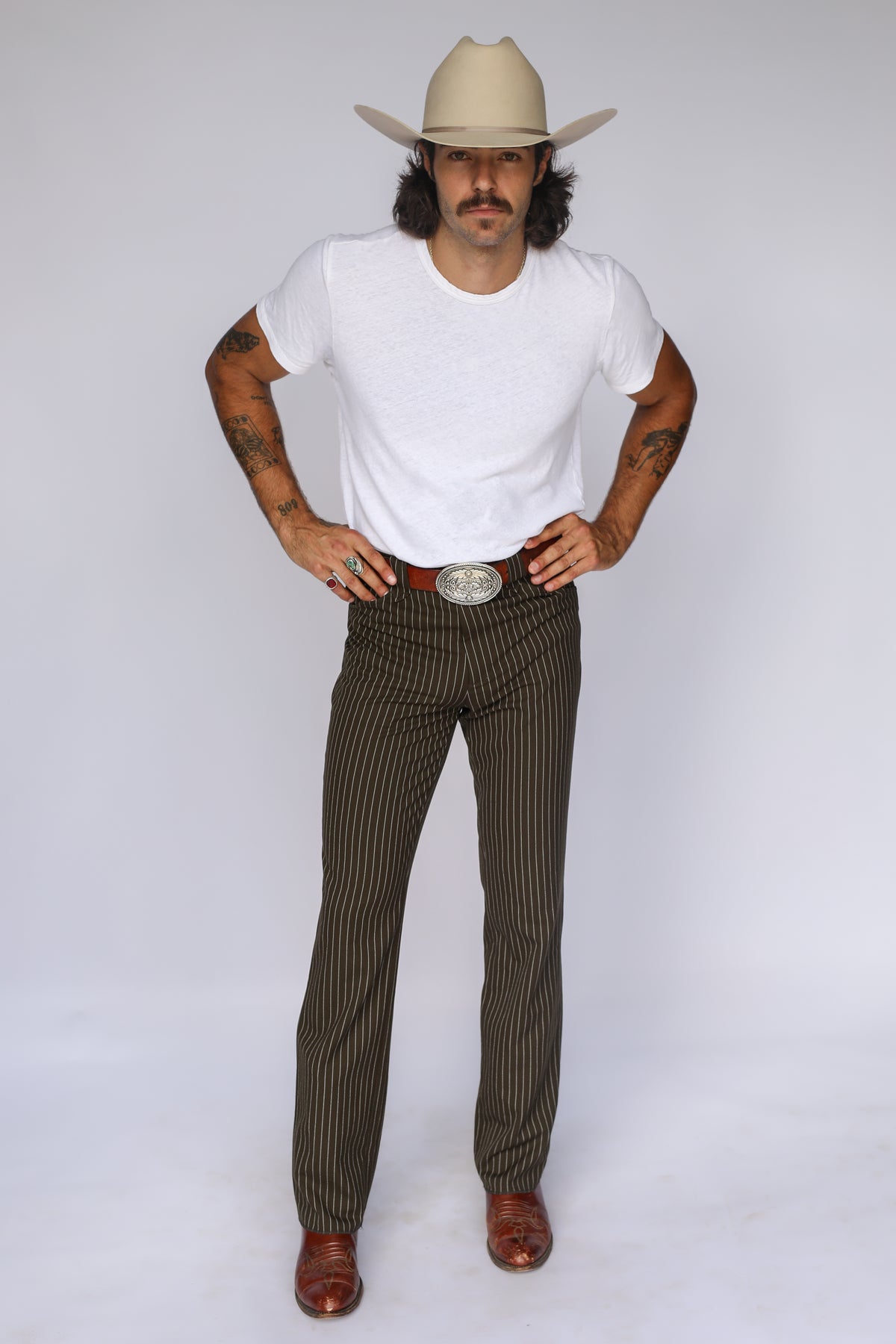 Man in a cream cowboy hat wearing the H Bar C Bakersfield brown pin stripe western pants with a white tee and silver buckle.