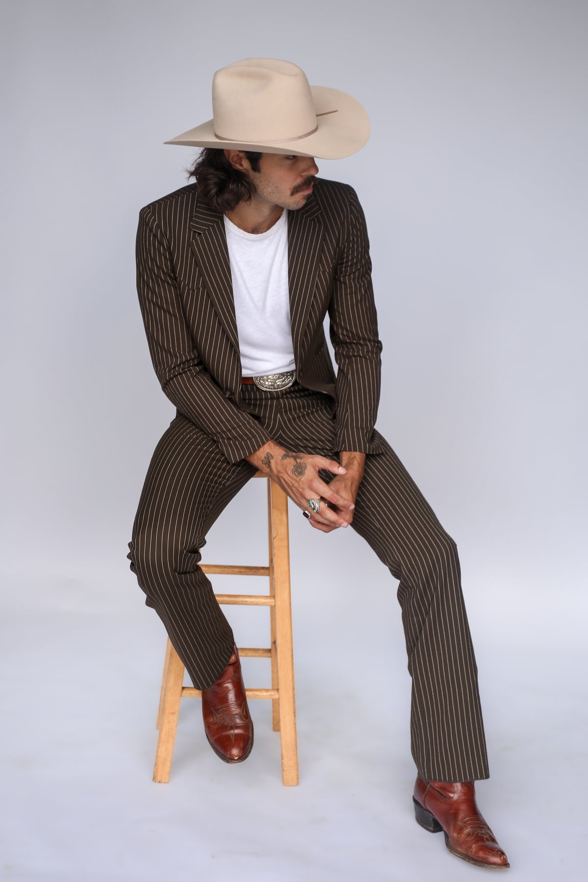 Man seated on a stool wearing the H Bar C Bakersfield brown pin stripe jacket and pants with a cowboy hat.