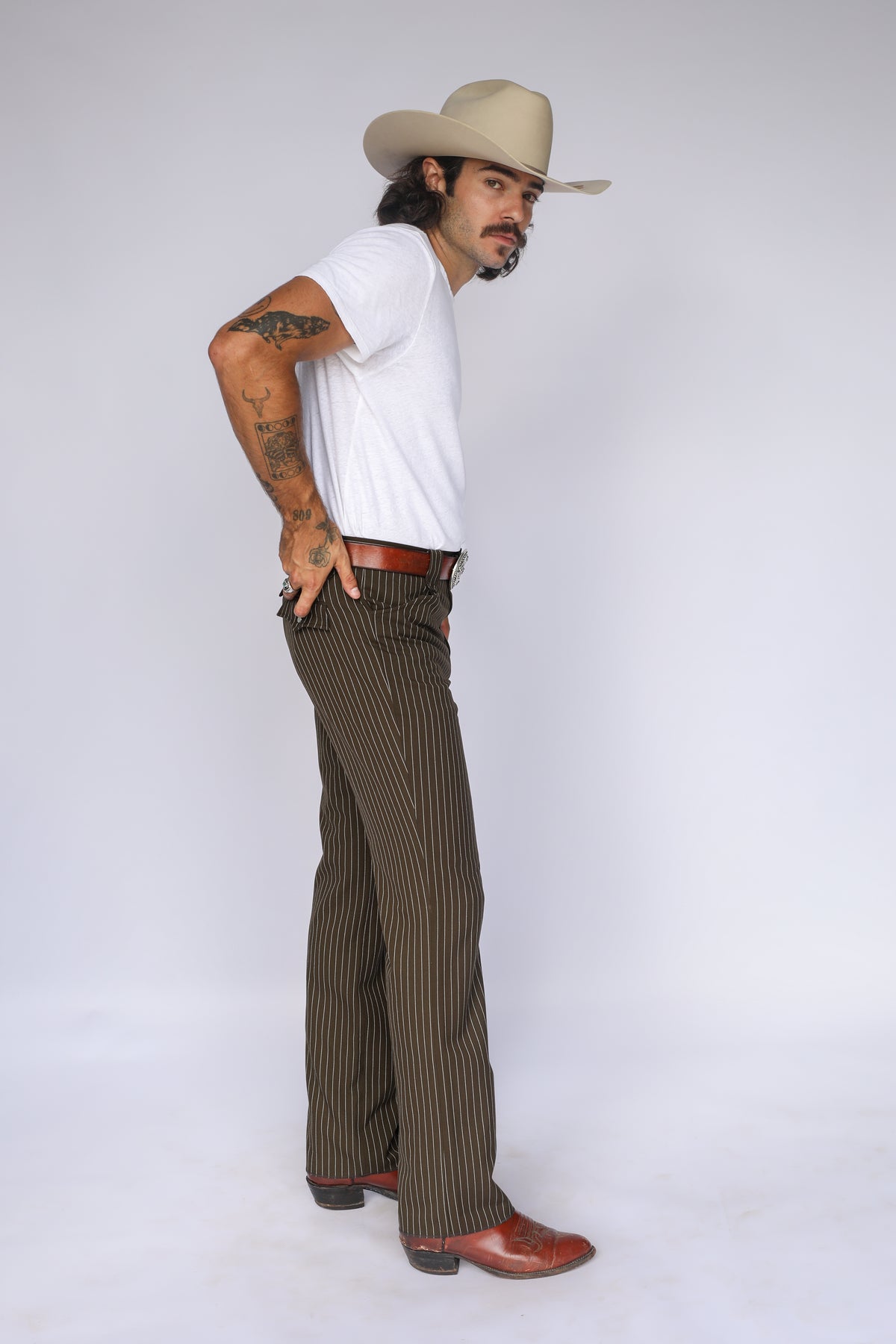 Man with hand on hip wearing the H Bar C Bakersfield brown pinstripe western pants, white tee, and cowboy hat.