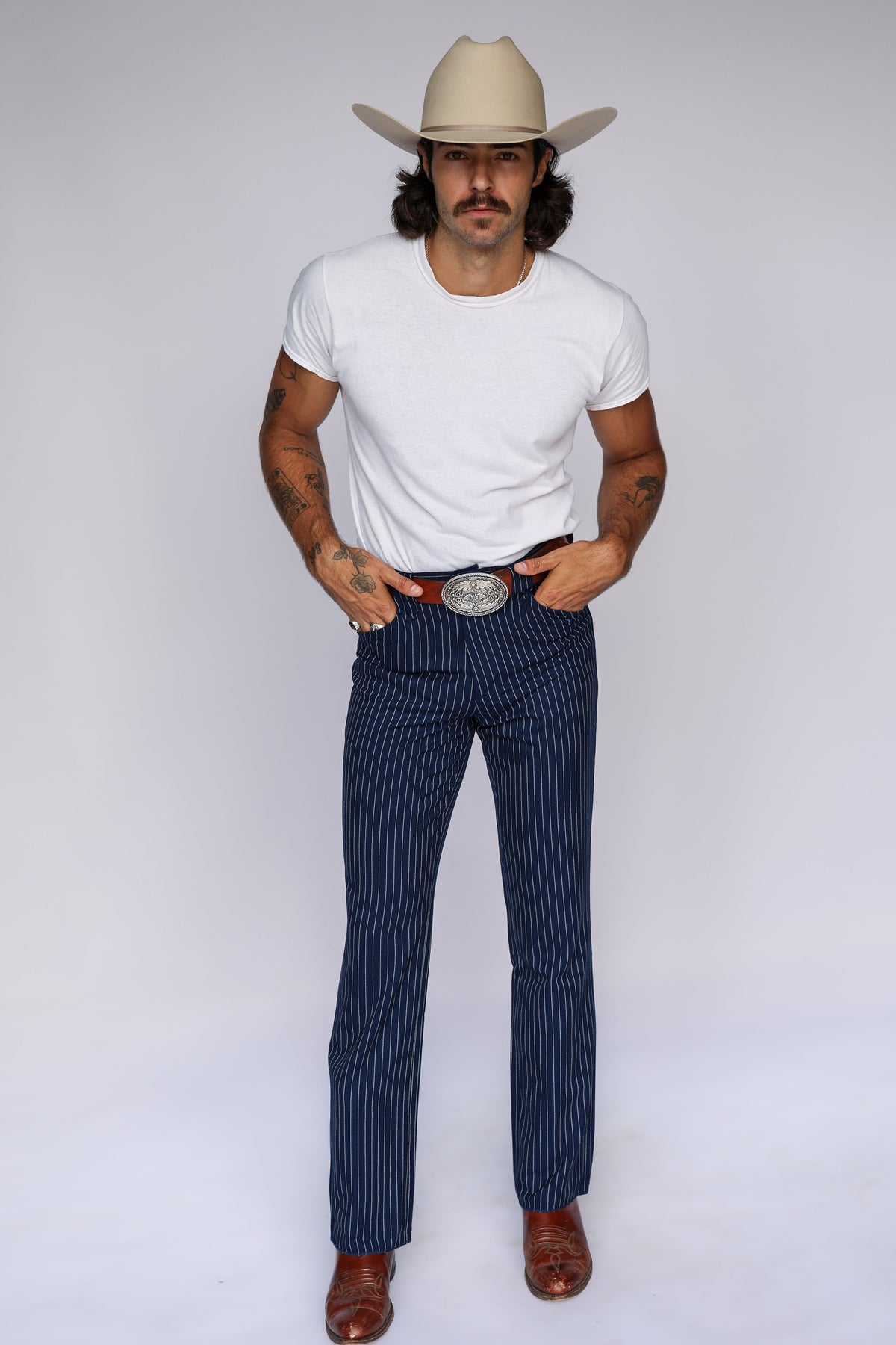 A man modeling a pair of navy blue pinstriped trousers with a large silver buckle, styled with a white t-shirt, brown boots, and a beige cowboy hat against a white studio background.