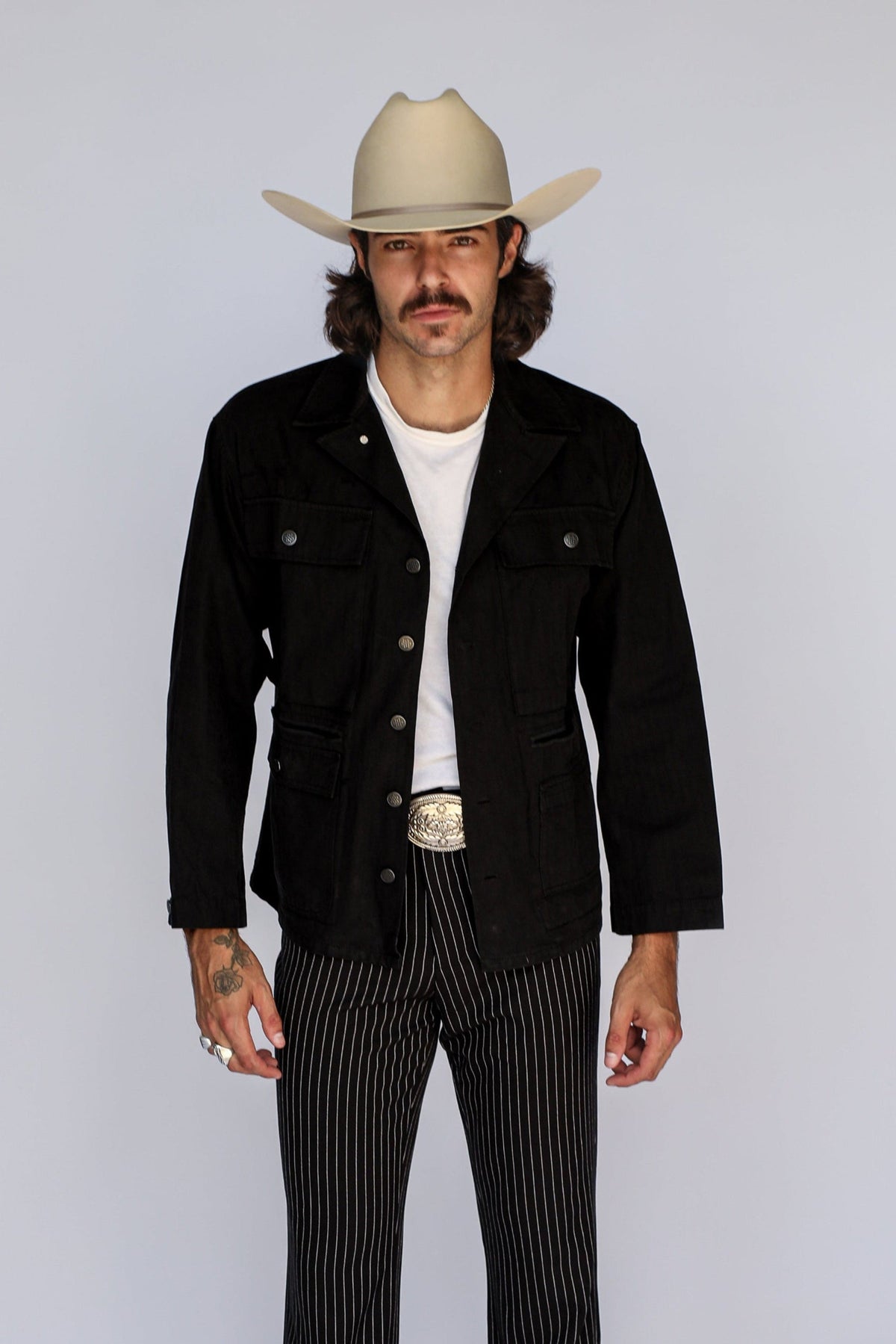 Man in a cowboy hat wearing the H Bar C Normandy chore coat in black, based on a 1940s WWII M-43, with pinstripe pants.