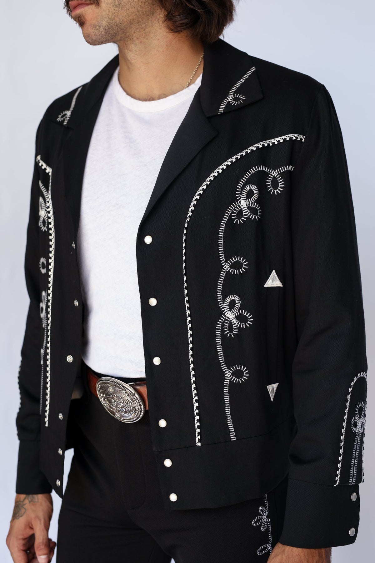 Front detail of the H Bar C Bolero men's black jacket showing white chain stitch embroidery, horseshoe appliqué, and a silver belt buckle.