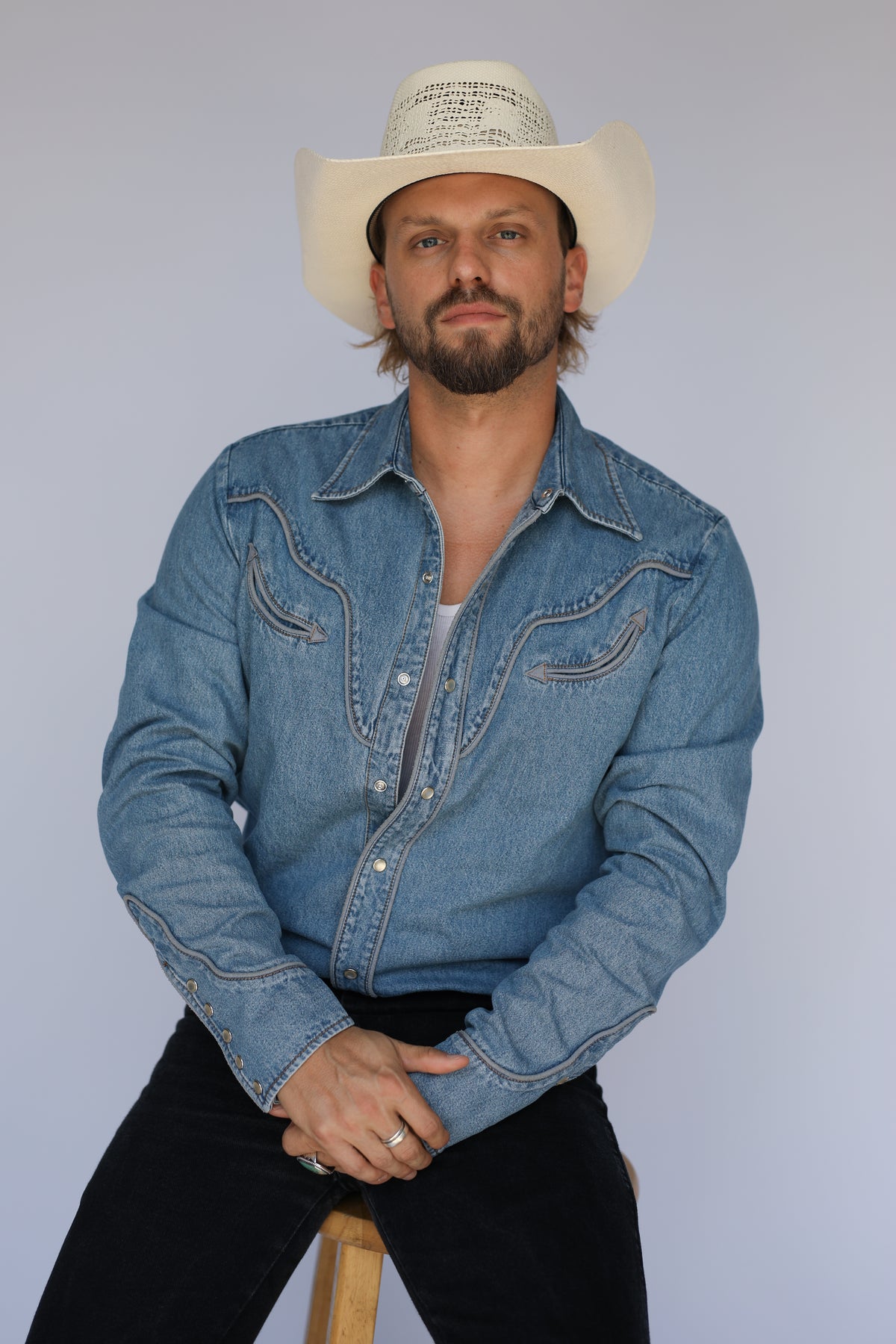 Man seated on a stool in a straw cowboy hat wearing the H Bar C San Fernando denim western shirt, a 100 year old H Bar C classic.