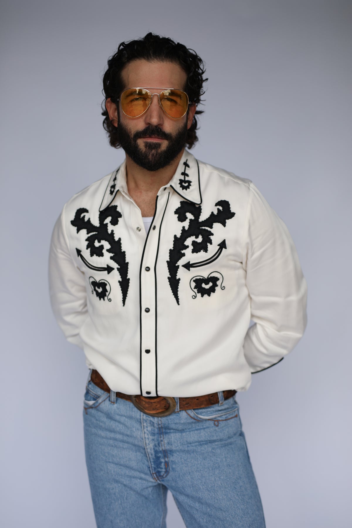 Man wearing the H Bar C Dali shirt in white with bold black flame embroidery, a tribute to Salvador Dalí, with light jeans.