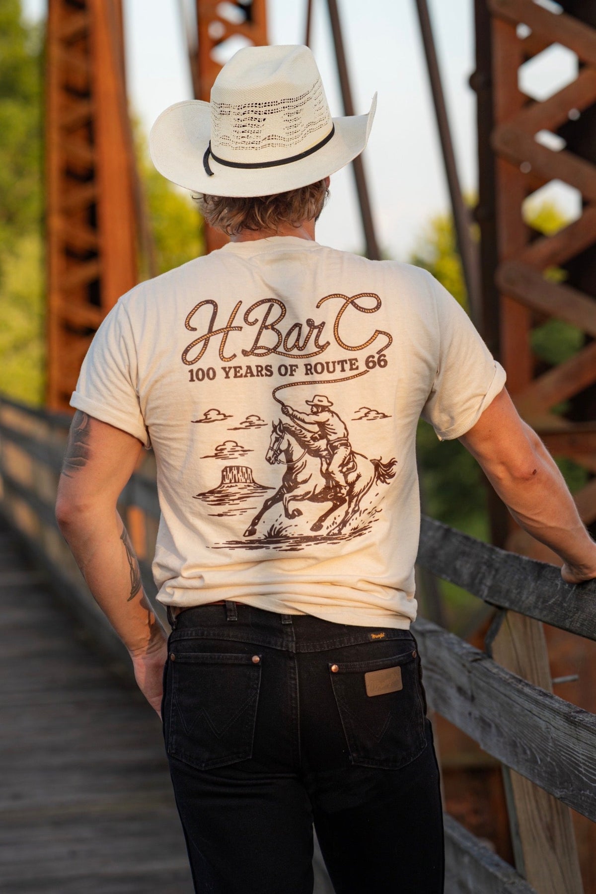 Back of the H Bar C Tulsa Rope T-shirt, a limited edition Route 66 Centennial tee with cowboy graphic, worn on a rusted iron bridge.