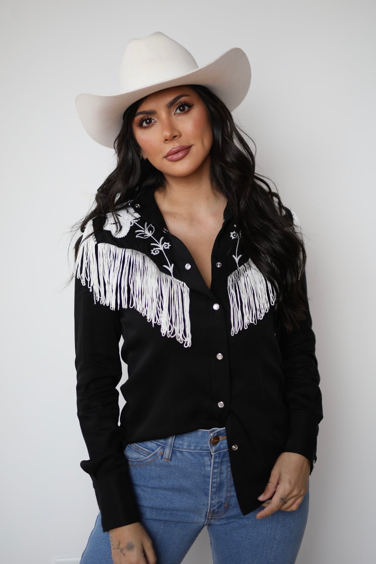 Woman in a cream cowboy hat wearing the H Bar C Taos women's shirt in black with white fringe and embroidery on the yoke, with jeans.