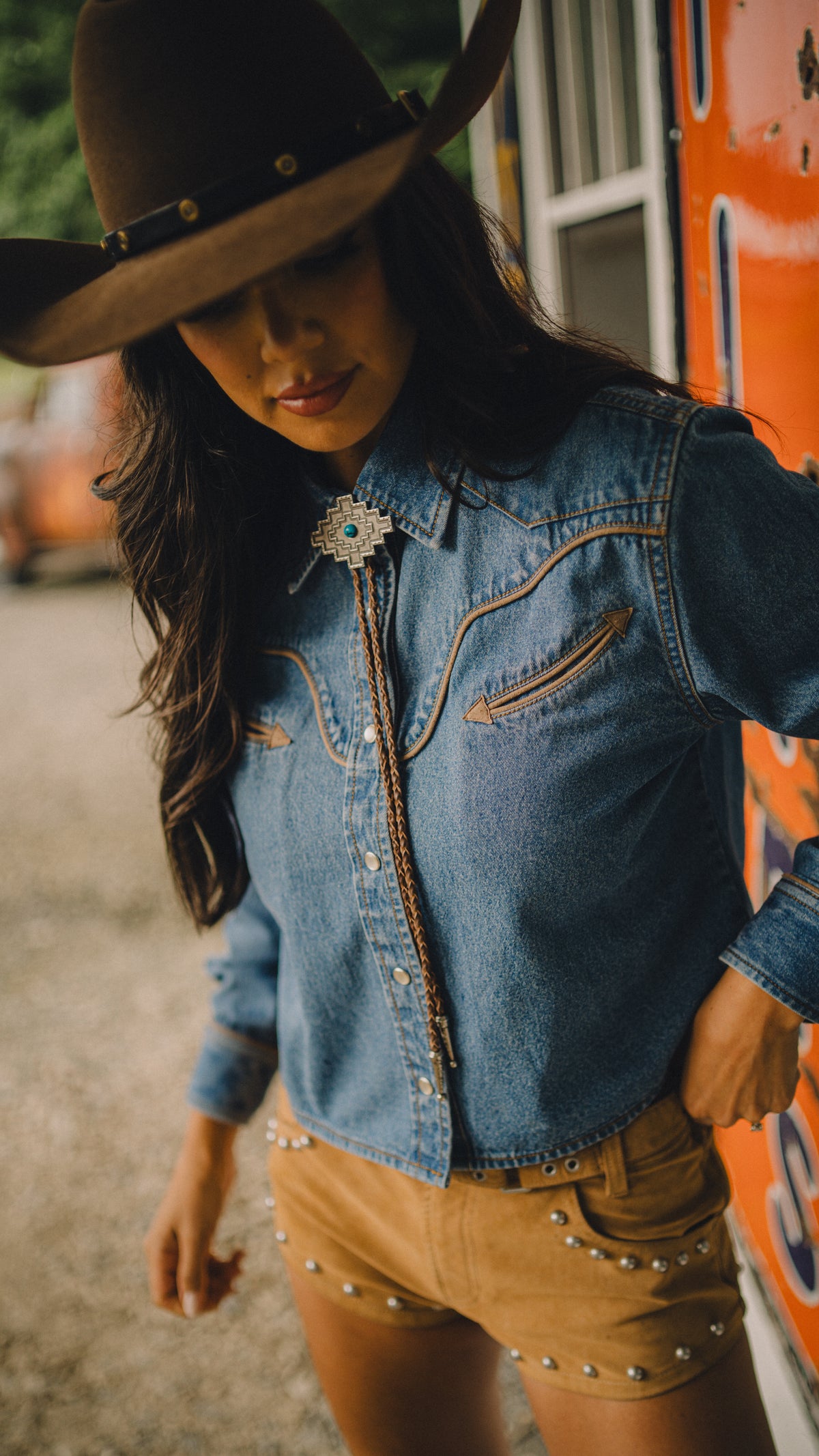 Woman in a cowboy hat wearing the Rustler x H Bar C denim cropped shirt with tan shorts outdoors.