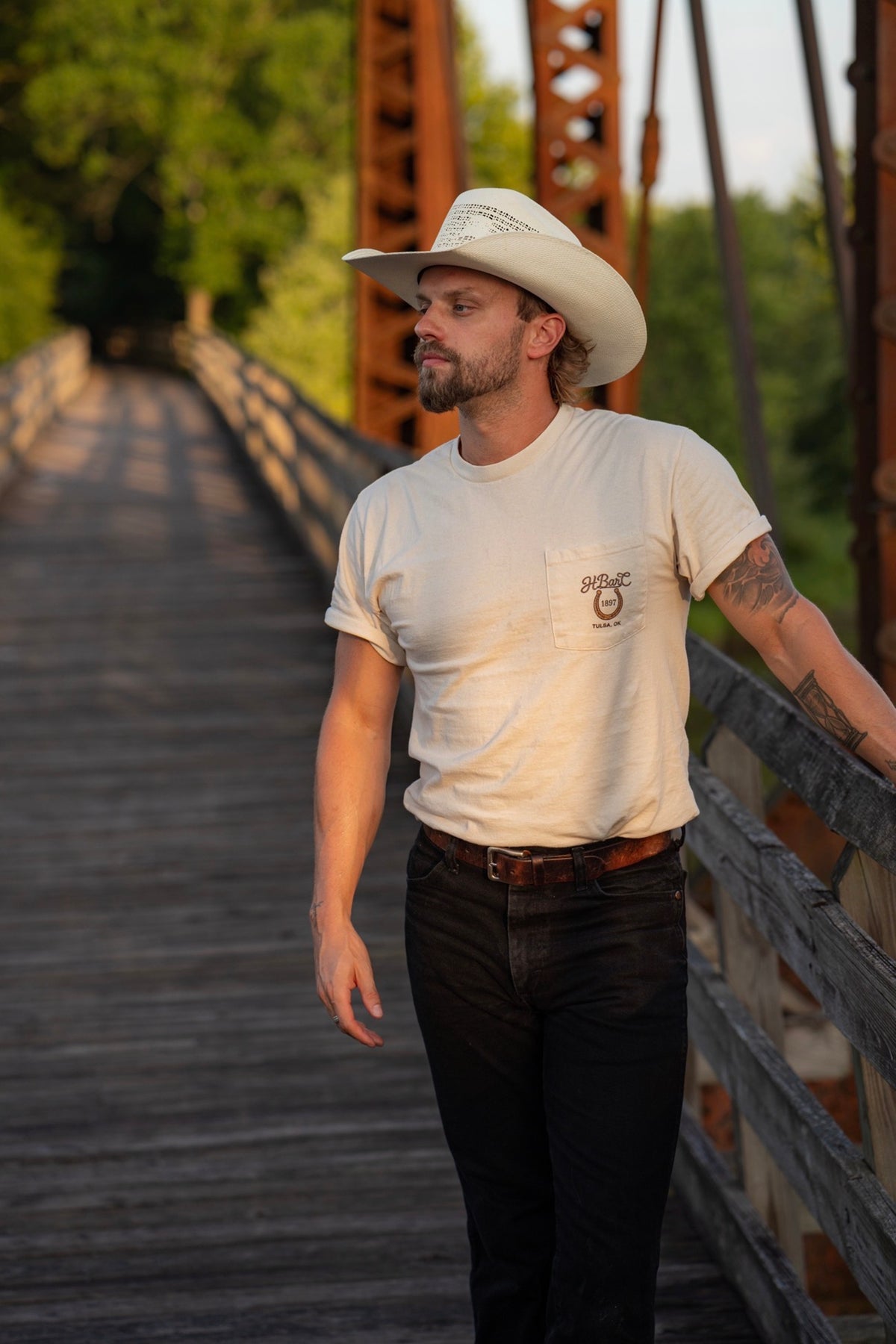 Front view of the cream H Bar C Tulsa Shield pocket tee on a man in a cowboy hat on a wooden bridge.