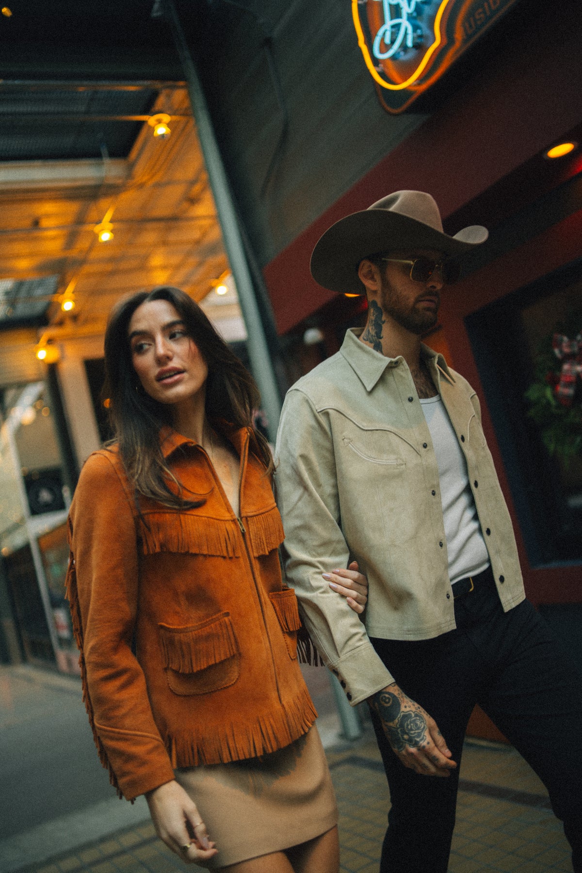 Couple on a neon lit street, the woman in the H Bar C Omaha rust suede fringe jacket.