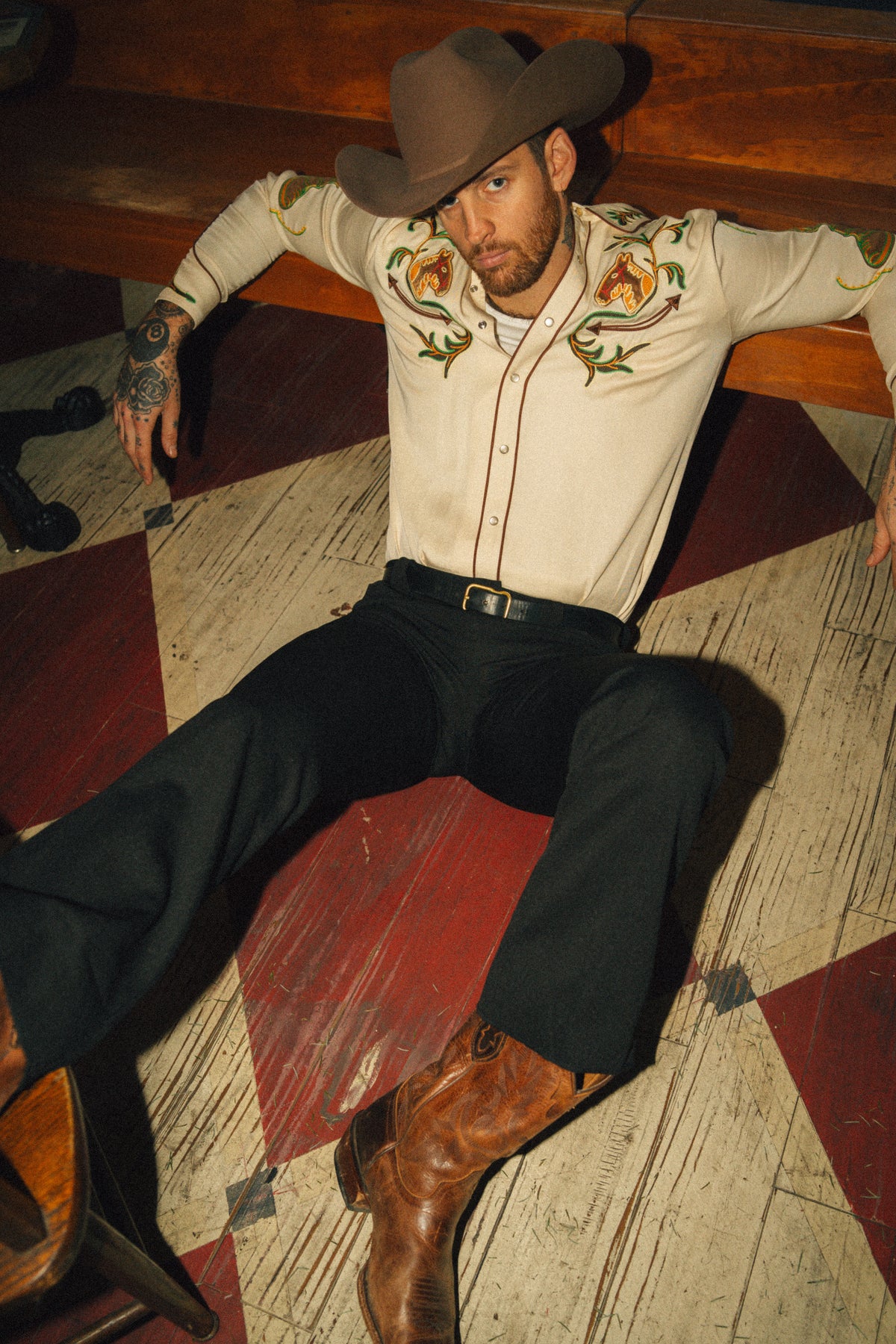 Man lying on a checkered floor wearing the H Bar C Calabasas off-white embroidered western shirt with black pants and boots.