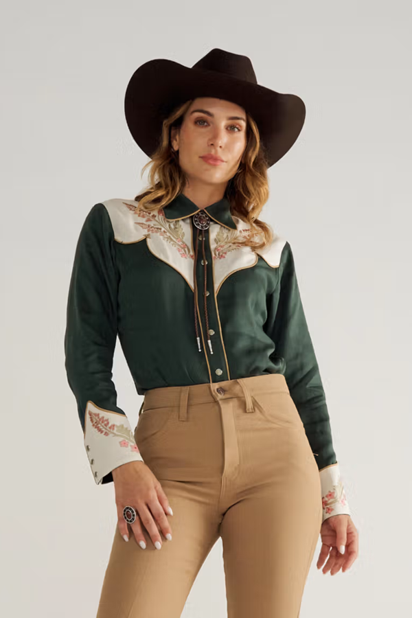 Woman in a cowboy hat wearing the Tecovas x H Bar C Birthday X shirt in hunter green with cream yokes and pink X embroidery.