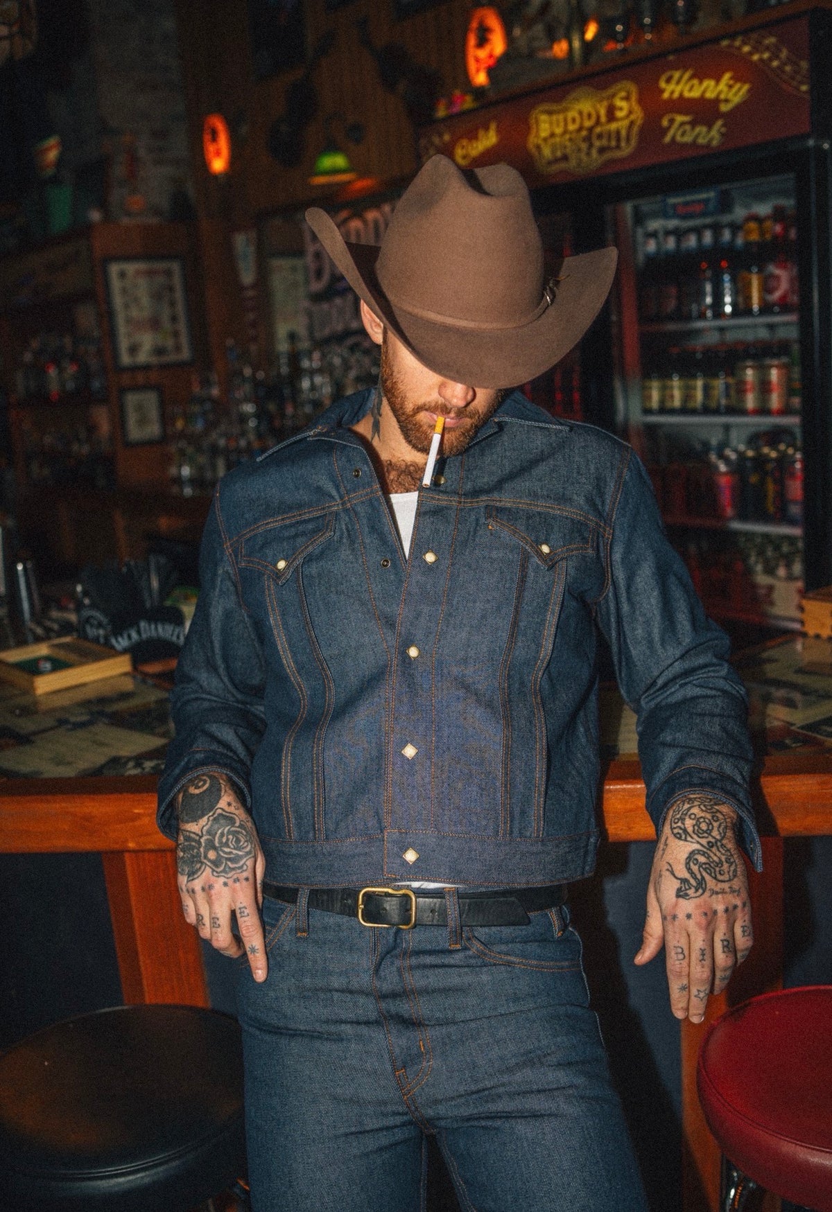 Man in a cowboy hat wearing the H Bar C Salina indigo denim jacket and jeans inside a neon-lit honky tonk bar.