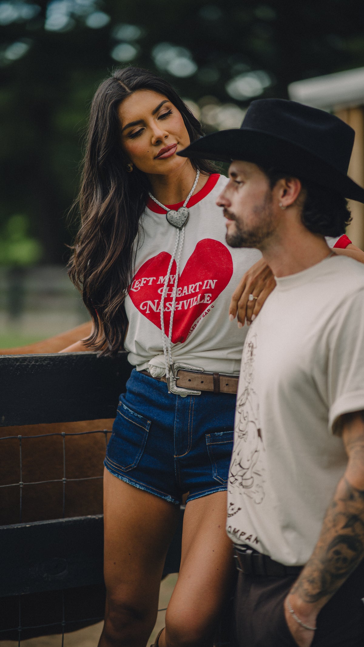 Couple at a wooden fence, woman wearing the H Bar C x Rustler Left My Heart in Nashville ringer and denim shorts.