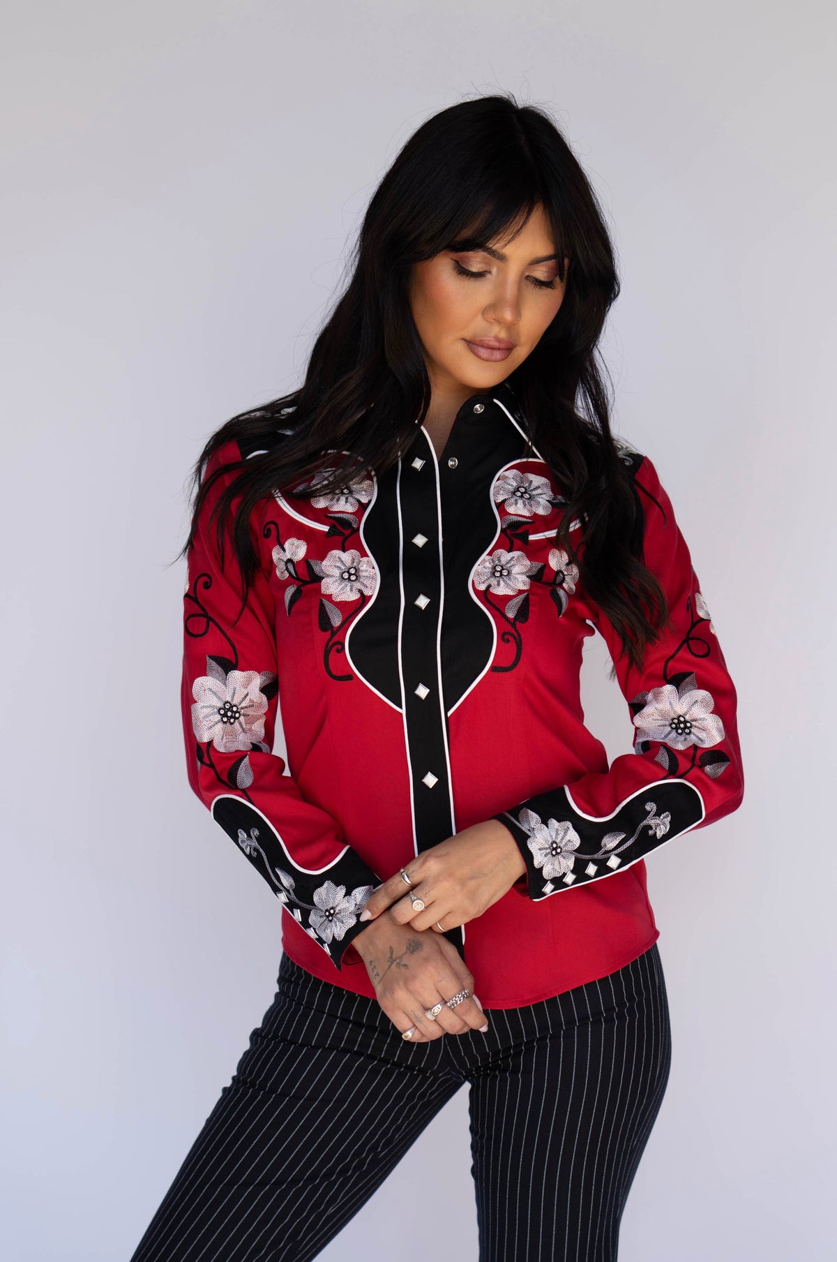 Woman wearing the H Bar C Santa Ana women's shirt in red with black yokes, white floral embroidery, and white piping, with pinstripe pants.