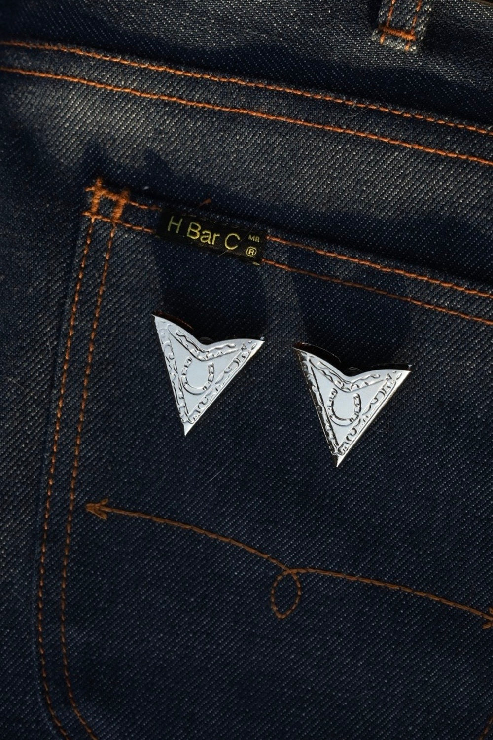 Pair of H Bar C engraved silver collar tips displayed on a dark denim back pocket.